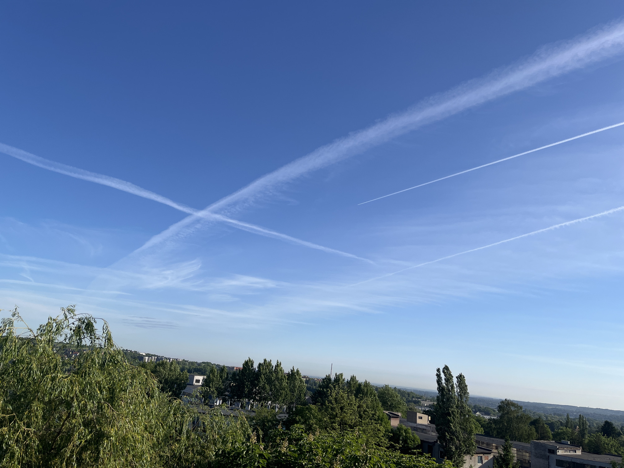 The image shows a daytime scene with a blue sky and multiple contrails from airplanes. In the foreground, there is greenery, including trees. Further back, buildings are visible amidst the trees, suggesting an urban or suburban area. The lighting appears to be bright and sunny. The location is Unknown, Unknown.