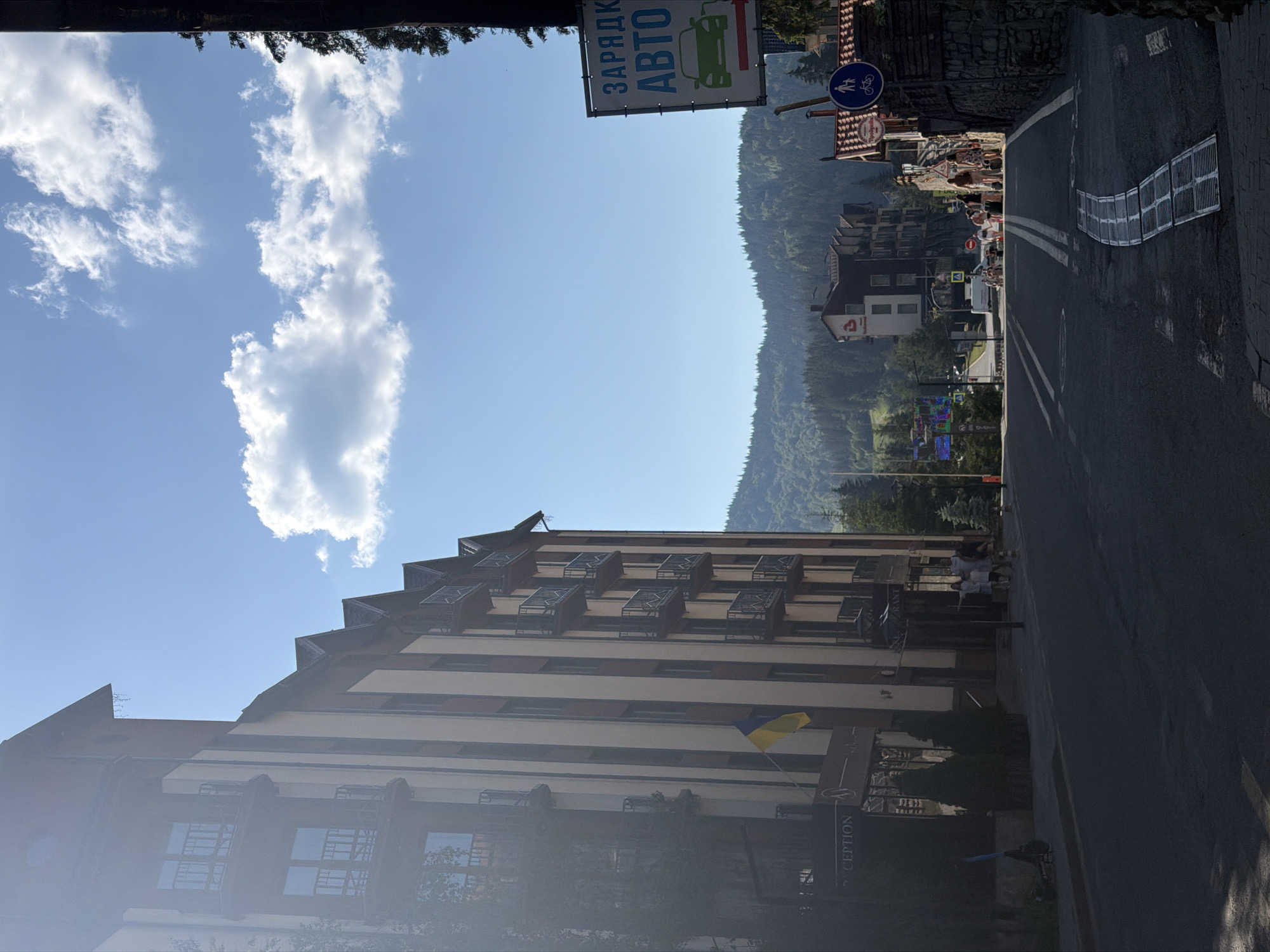 The image depicts a street scene in Polianytsia, Ukraine, likely in a resort area, with a large hotel on the left and a road leading into a hilly, forested landscape. The hotel is a multi-story building with balconies and a Ukrainian flag prominently displayed. Several people are walking on the sidewalk in front of the hotel, and a scooter is parked nearby. Across the street, there is a sign for "CHARGING AUTO" indicating an electric vehicle charging station. Other buildings, including a smaller hotel or guesthouse, are visible further down the road, nestled against the backdrop of dense green hills covered in pine trees. The sky is blue with scattered clouds, and the sunlight suggests it is daytime, possibly late morning or early afternoon, given the shadows. The road has clear lane markings and a storm drain cover in the foreground.