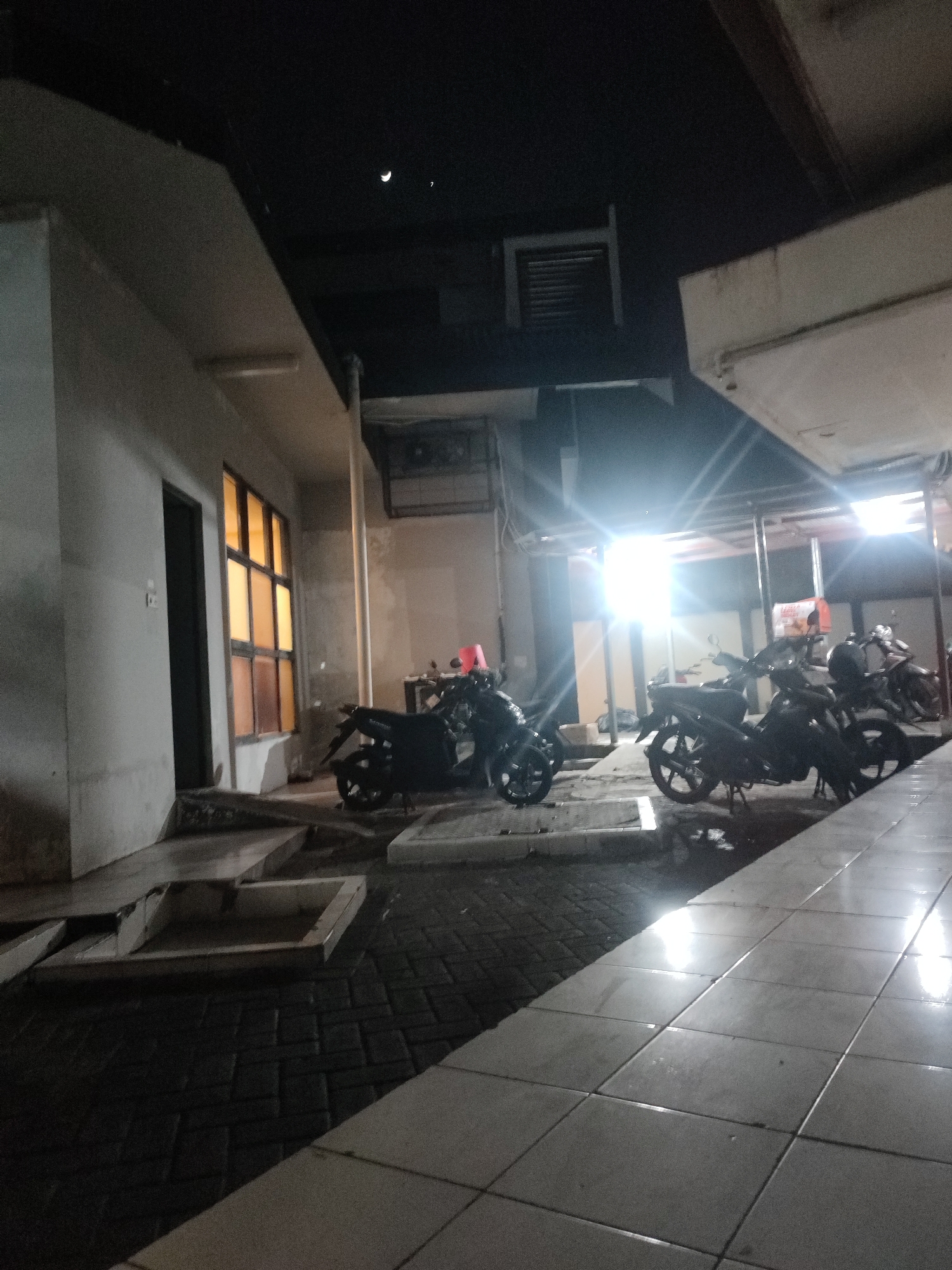 The image depicts an outdoor scene at night, likely in Jember, Indonesia. The foreground is dominated by a tiled walkway, wet and reflecting the bright overhead lights. To the left, a building with a textured, pale facade is visible, featuring a dark doorway and a window with multiple panes illuminated by a warm, orange light. A series of concrete steps leads down from the building's entrance. Beyond the building, a paved area with brick-like patterns is observed, with several dark-colored motorcycles parked. One motorcycle in the mid-ground is particularly prominent, with a red box or container visible on its rear. In the background, bright overhead lights cast strong glares, and what appears to be a metal grate is set into the dark ceiling of an overhang. The sky above is dark, with a crescent moon and another faint celestial body visible. The overall impression is of a dimly lit, utilitarian area, possibly a parking space or service entrance associated with a building. There are no people visible in the image.