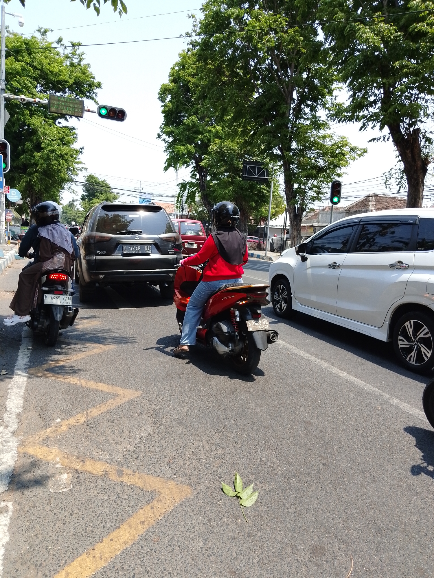 In Pasuruan, Indonesia, on a clear, sunny day, traffic is stopped at an intersection. A traffic light is visible, showing a green signal. Several vehicles are present, including a black Mitsubishi Pajero Sport SUV in the center, a white car on the right, and two motorbikes with riders. The rider on the left motorbike wears a dark brown outfit and a black helmet. The rider on the right motorbike is wearing a red top and blue jeans, also with a black helmet. The road has yellow zig-zag markings on the asphalt, indicating a no-parking zone. Lush green trees line the street, and buildings can be seen in the background.