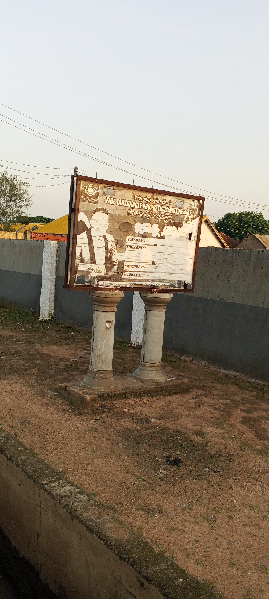 A weathered billboard stands on two ornate pedestals in a dirt lot. The billboard advertises "FIRE TABERNACLE PROPHETIC MINISTRIES INT (Home Of Solution)". A faded image of a man in a suit is on the left side of the billboard. To the right, the days of the week, including "TUESDAY'S", "THURSDAY'S", "SATURDAY'S", and "SUNDAY'S", are listed, but much of the text is obscured by damage to the billboard. The background shows a wall, a yellow-roofed building, and some trees. The sky is clear and pale. This scene is located in the city of Jalingo, Nigeria, where the billboard is a prominent fixture in the community.