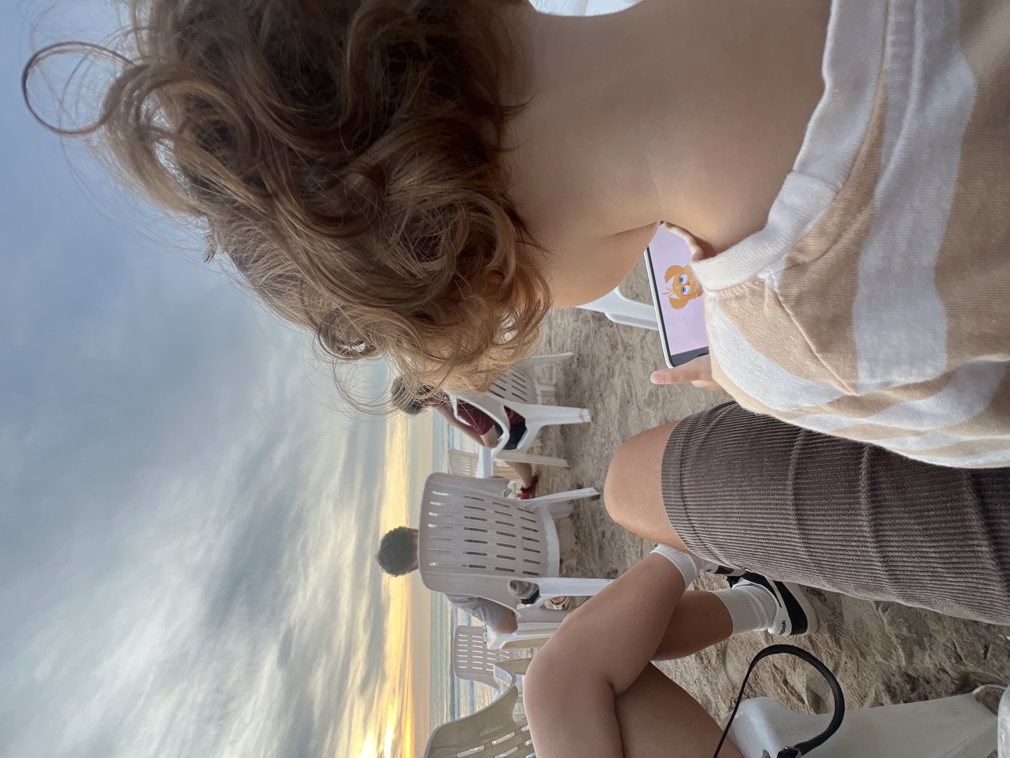 This image captures a beach scene in Choeng Thale, Thailand, with a focus on a young child holding a tablet, viewed from behind. The child has curly brown hair and is wearing a striped t-shirt. In the foreground, the legs of at least two individuals are visible, seated on the sand. One person is wearing brown corduroy shorts and white socks with black sneakers. Another person's legs are also visible, wearing a white sock. In the background, several white plastic chairs are arranged on the sandy beach. Three people are seated in these chairs, facing away from the camera towards the ocean. The sky above is cloudy with hints of sunset colors. The ocean is visible in the distance. The overall atmosphere suggests a relaxed evening on the beach.