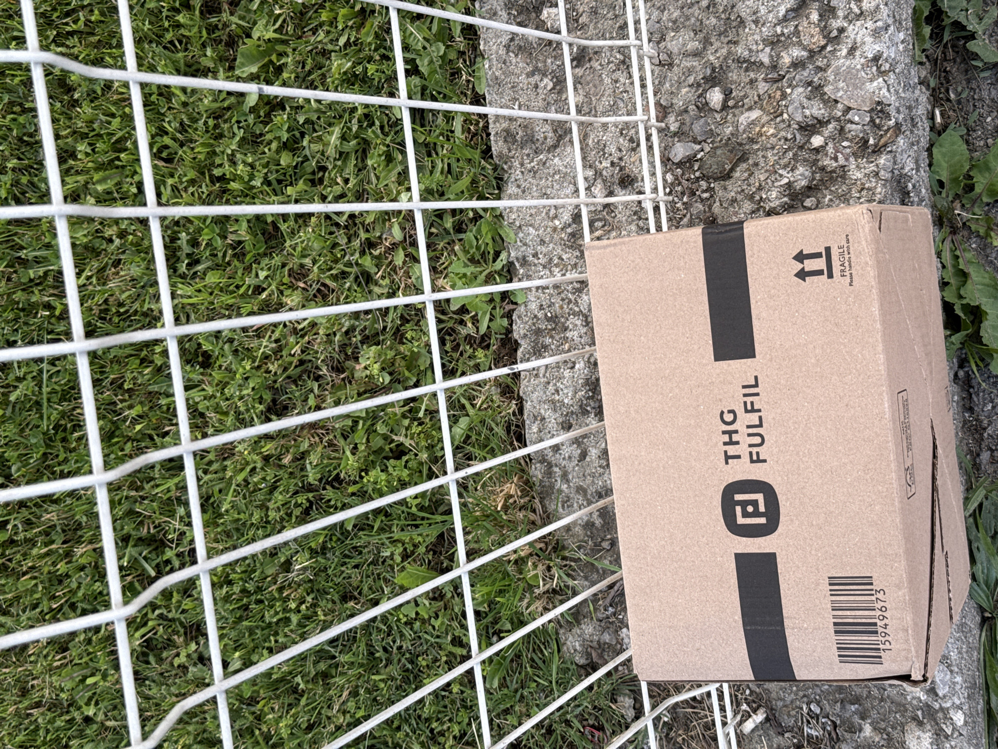 A cardboard box labeled "THG FULFIL" with a barcode and a "Fragile" warning is situated on a concrete surface near a white metal grid, partially extending onto a grassy area. The scene appears to be outdoors. There is no discernible evidence of it being in Sofia, Bulgaria. There are no people visible in the image.