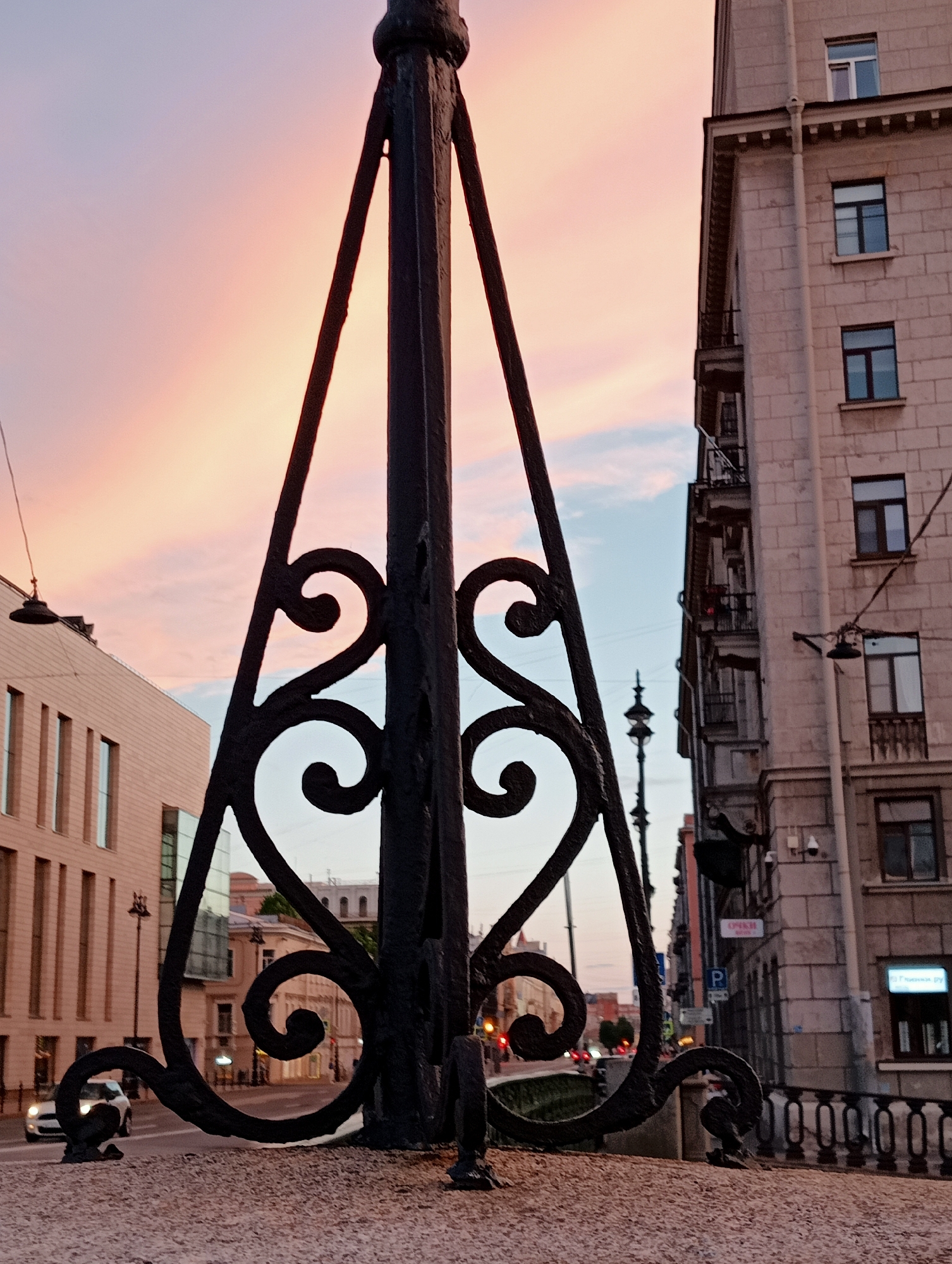The image shows a view of a street in Saint Petersburg, Russia, at sunset. In the foreground, a decorative black metal lamppost with ornate scrollwork is prominently displayed. Behind the lamppost, a light-colored building with stone facade and balconies rises on the right, while another modern building with large windows is visible on the left. The sky displays a soft gradient of pink, orange, and blue, indicative of twilight. A street with a few cars and streetlights is visible in the background, suggesting an urban setting. The overall atmosphere is serene and evocative of a European city at dusk.