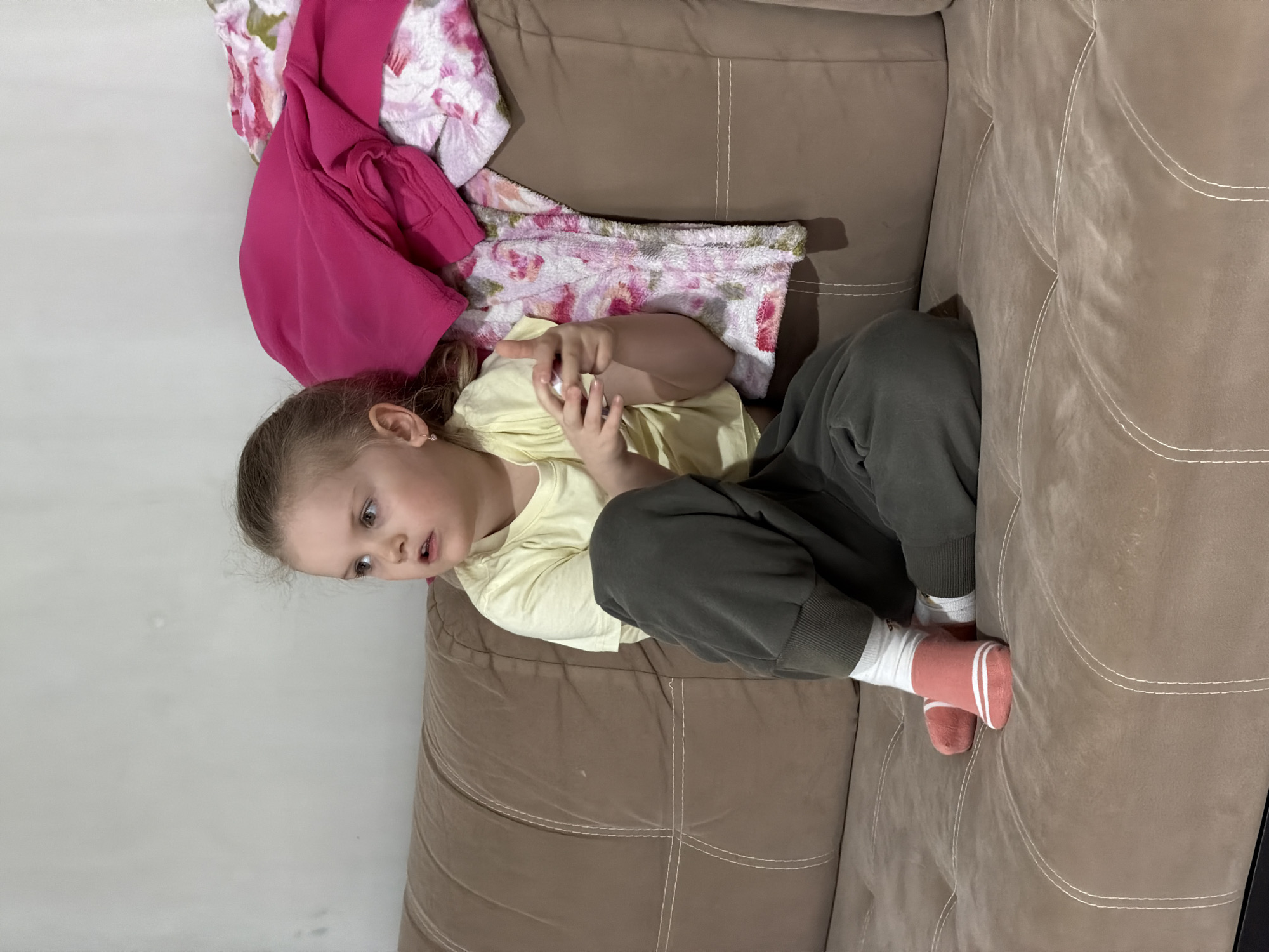 A young child lies on a brown upholstered sofa. She wears a yellow short-sleeved shirt, dark green sweatpants, and pink socks with white stripes. Her hair is light brown, pulled back from her face. A pink shirt and a floral blanket are near her head on the sofa. The scene is inside, with a light-colored wall visible in the background. There is no indication of the location being Chaikovsky, Russia.