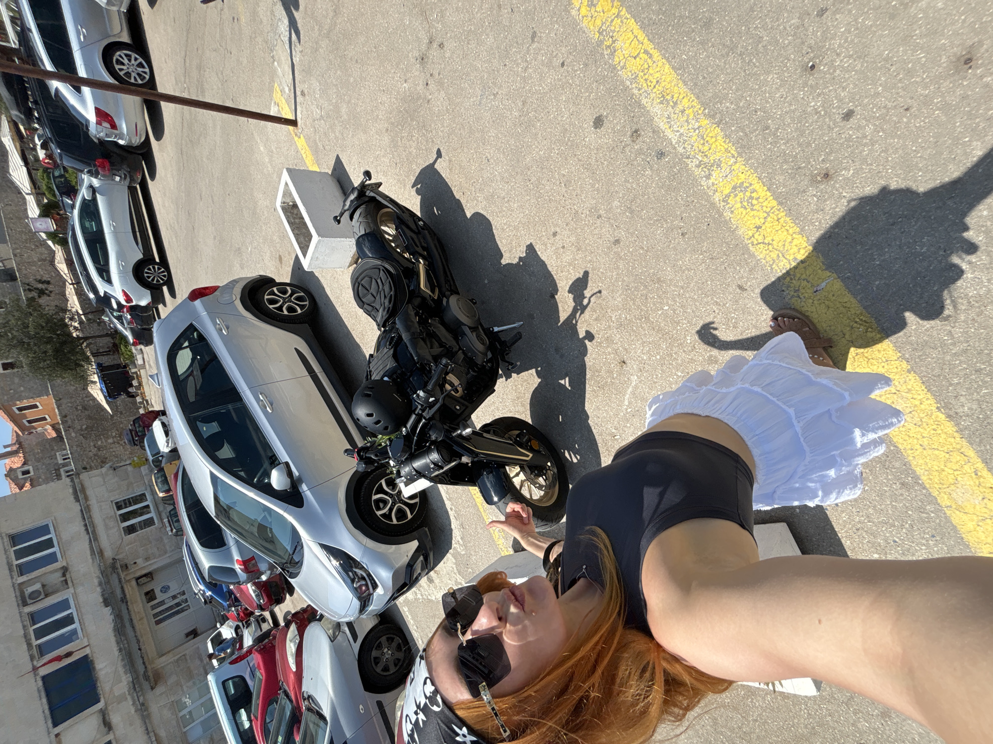 A woman with long red hair, wearing a black tank top, a white ruffled skirt, sunglasses, and a bandana, is taking a selfie in a parking lot. She is making a kissing face and giving a thumbs up. Next to her is a black motorcycle with a helmet on its seat. The background shows several parked cars, including a silver Kia Picanto and a white SUV. The location appears to be a public parking area, likely in a city given the surrounding buildings and the presence of other vehicles. The scene is illuminated by bright sunlight, casting shadows on the asphalt. The ground is marked with yellow lines, indicating parking spaces. The buildings in the background have a Mediterranean architectural style, with stone walls and red-tiled roofs, suggesting a location in southern Europe. The presence of a white concrete planter box adds to the urban setting. The overall atmosphere is casual and summery.