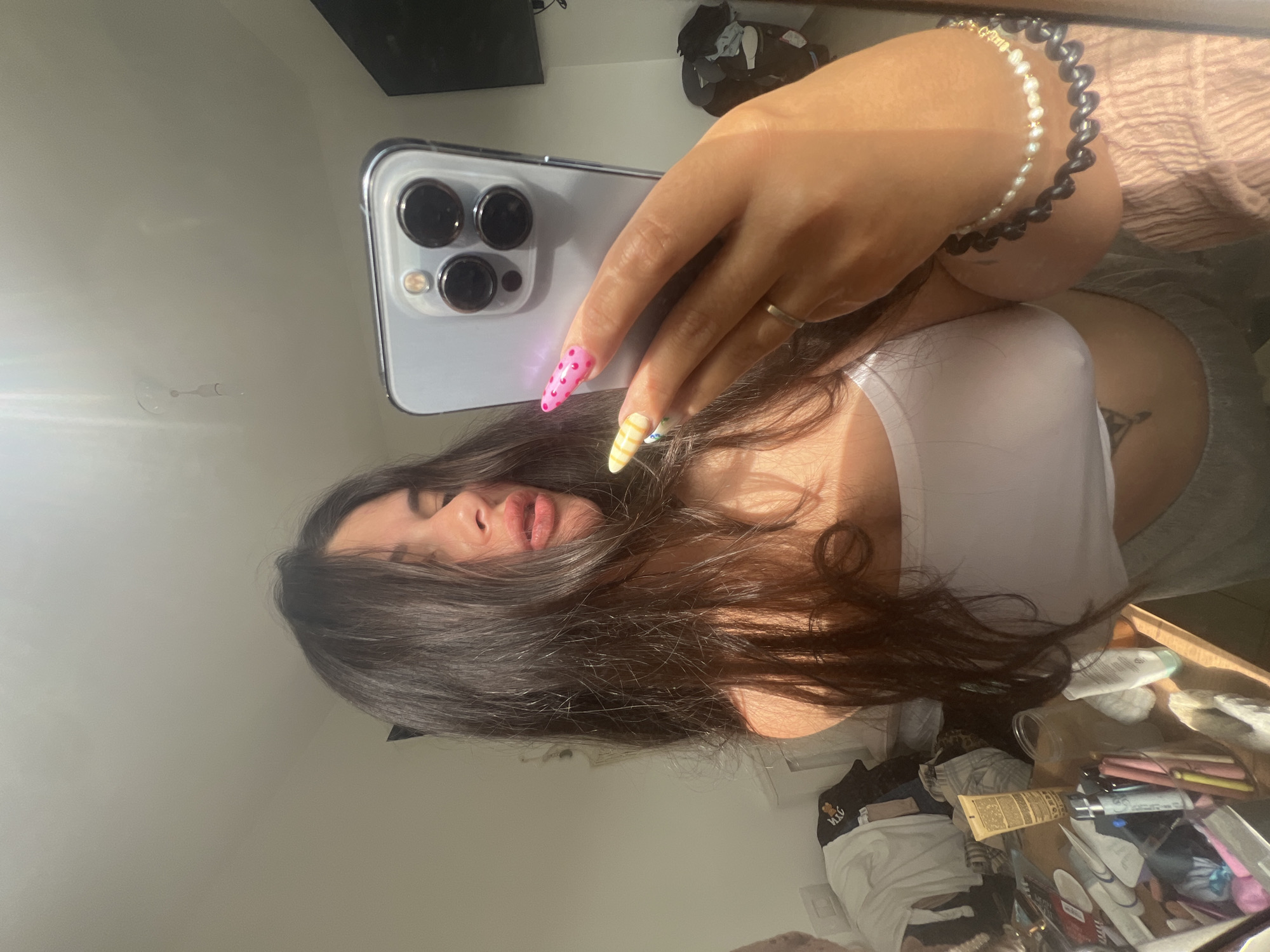 A woman is taking a selfie in an indoor setting. She is holding a light blue smartphone with three camera lenses. The woman has long, dark hair and wears a white tank top. A gray garment is also visible. She has two bracelets on her wrist and her fingernails are painted in different patterns. The background shows a cluttered table with various objects including cosmetics, tubes, and clothing. The specific location within Kiryat Bialik, Israel is not discernible from the image.