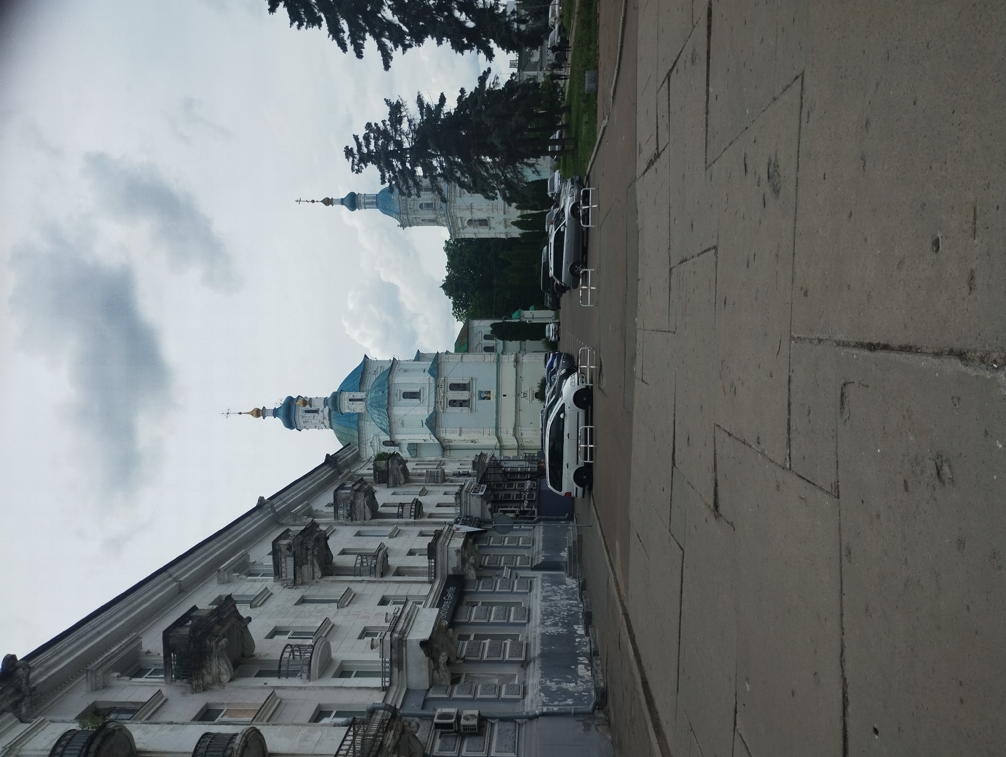 This is a medium shot of a street in Sumy, Ukraine, with an old apartment building on the left and two white churches with blue domes on the right. The apartment building has multiple balconies with wrought-iron railings. The churches are ornate with architectural details. Several cars are parked in a lot in front of the churches, and some people are visible in the distance. The pavement in the foreground is made of large concrete slabs. The sky is overcast. The scene appears to be captured during the day.