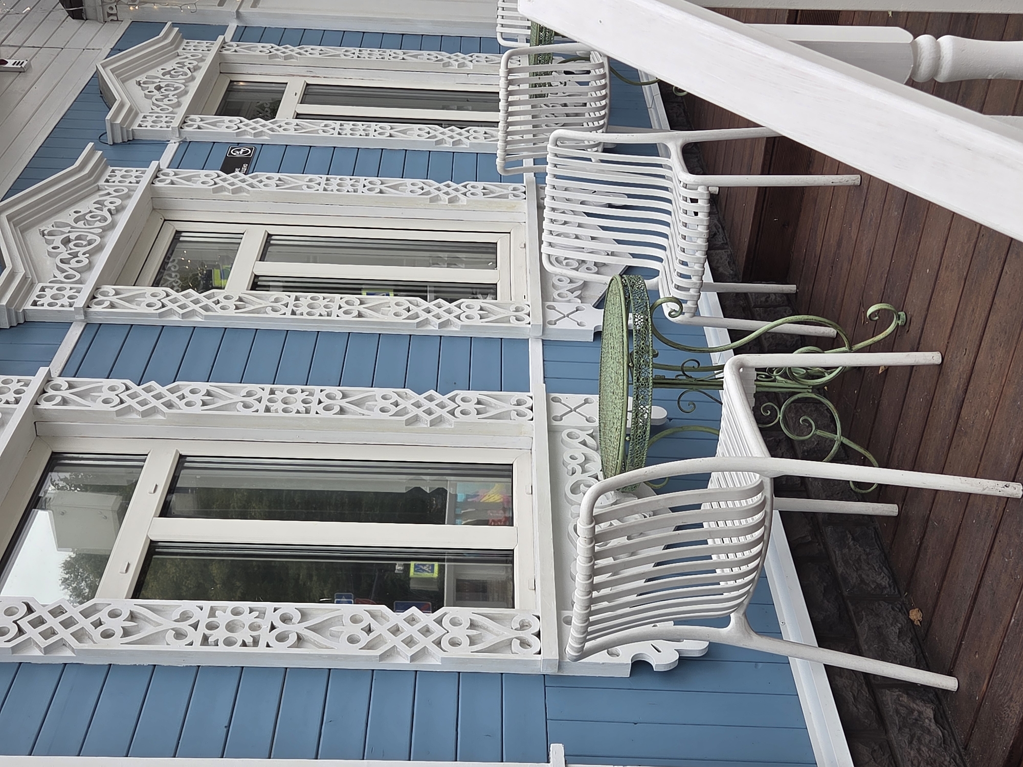 The image shows a close-up of a wooden porch in Zvenigorod, Russia, featuring a light blue wooden facade with white carved wooden trim around three windows. Two white metal chairs with slatted seats and backs are positioned on the porch. In front of one chair, a green metal round table with a decorative base is visible. The porch floor is made of dark brown wooden planks. The overall scene depicts an outdoor seating area of a building with traditional Russian wooden architecture.
