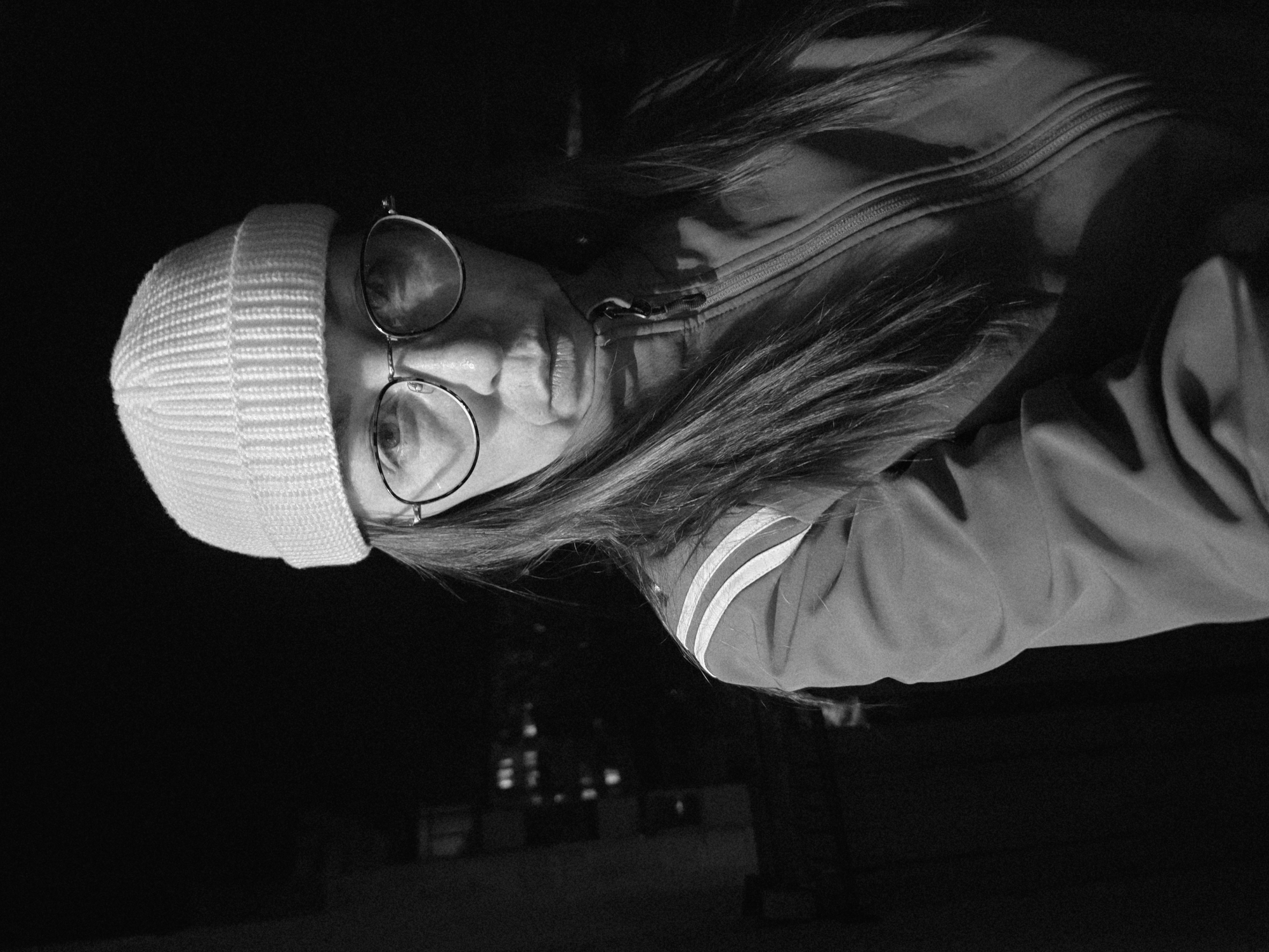 A monochrome, chest-up portrait shows an individual facing forward, staring directly into the camera. The person wears a light-colored, ribbed knit beanie and round-rimmed spectacles. Long, dark hair extends from under the beanie, down past the shoulders. The individual is clothed in a dark, zippered jacket, partially zipped, with two prominent light-colored horizontal stripes visible on the left shoulder. The lighting is low-key, with the subject illuminated from the front, casting subtle shadows. The background is predominantly dark, with indistinct, blurred lights visible in the distance to the left, characteristic of an urban night scene. The location is Chaikovsky, Russia.