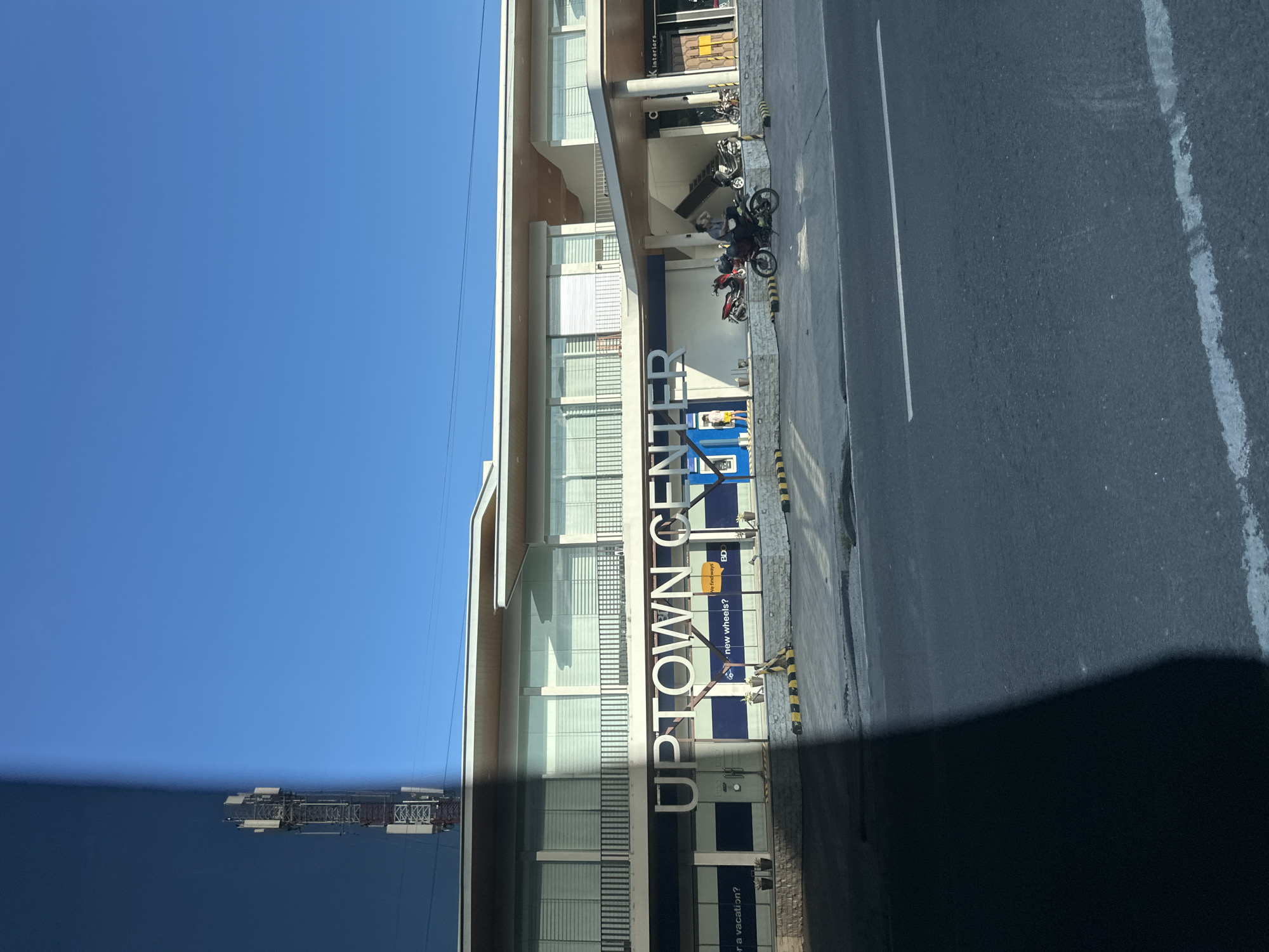 This daytime photograph captures the exterior of the "Uptown Center" commercial building from across an asphalt road in Davao City, Philippines. The left vertical third of the image is obscured by a sharp, dark shadow, suggesting a strong light source from the right relative to the photographer's position.
The "Uptown Center" is a multi-level building with a modern design, featuring a light-colored facade and a distinctive flat roof with an overhang. The upper level incorporates large windows with horizontal blinds or louvers and a dark protective railing. The building's name, "UPTOWN CENTER," is prominently displayed in large, white, sans-serif capital letters on the lower level facade. Below this, various commercial signage is visible, including phrases like "...or a vacation?", "new wheels?", "we find ways," and "BD." On the far right, a sign indicating "K interiors" is present above an entrance with a wooden door. A blue machine, resembling an ATM, is located centrally on the ground level. Potted plants are arrayed in front of the lower-level windows, and yellow and black striped concrete barriers delineate the edge of the building's parking or pedestrian area.
Several motorcycles are present in front of the building. A male individual, dressed in a dark shirt and pants, is seated on a black motorcycle near the building's covered entryway on the right side. A red motorcycle is parked nearby. Another person wearing yellow shorts is partially visible near the blue machine. Additional individuals are partially discernible near the "K interiors" entrance.
The foreground features an asphalt road with visible white lane markings; a solid white line runs horizontally across the mid-ground, and a faded wider white line is present closer to the bottom edge. In the upper left background, partially obscured by the dark shadow, a tall, metal communication tower equipped with multiple antenna arrays stands against a clear, vibrant blue sky, indicating fair weather.