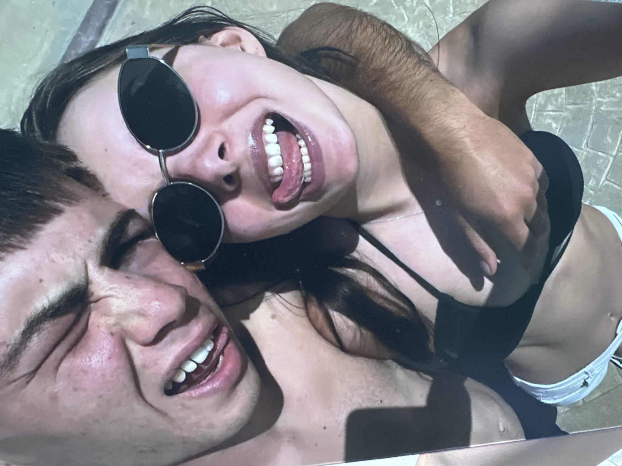 In this close-up photograph, a young couple is captured in what appears to be an outdoor setting with a tiled surface visible in the background, suggestive of a poolside or patio area. The focus is on their faces and upper bodies, with the lighting casting shadows that highlight their features.
On the left, a young male is seen with his eyes squeezed shut and his mouth open, revealing teeth and tongue. His skin shows a scattering of freckles. He is shirtless, and his right arm is around the female.
On the right, a young female is wearing oversized, oval-shaped sunglasses. Her mouth is open, with her tongue extended outwards. She has on a black bikini top and white bikini bottoms. Her left arm is around the male's neck.
The image is taken from a high-angle perspective, looking down at the couple. The overall impression is of a candid, playful moment shared between two individuals. There is no specific indication of the location being in the city of Sofia, Bulgaria.