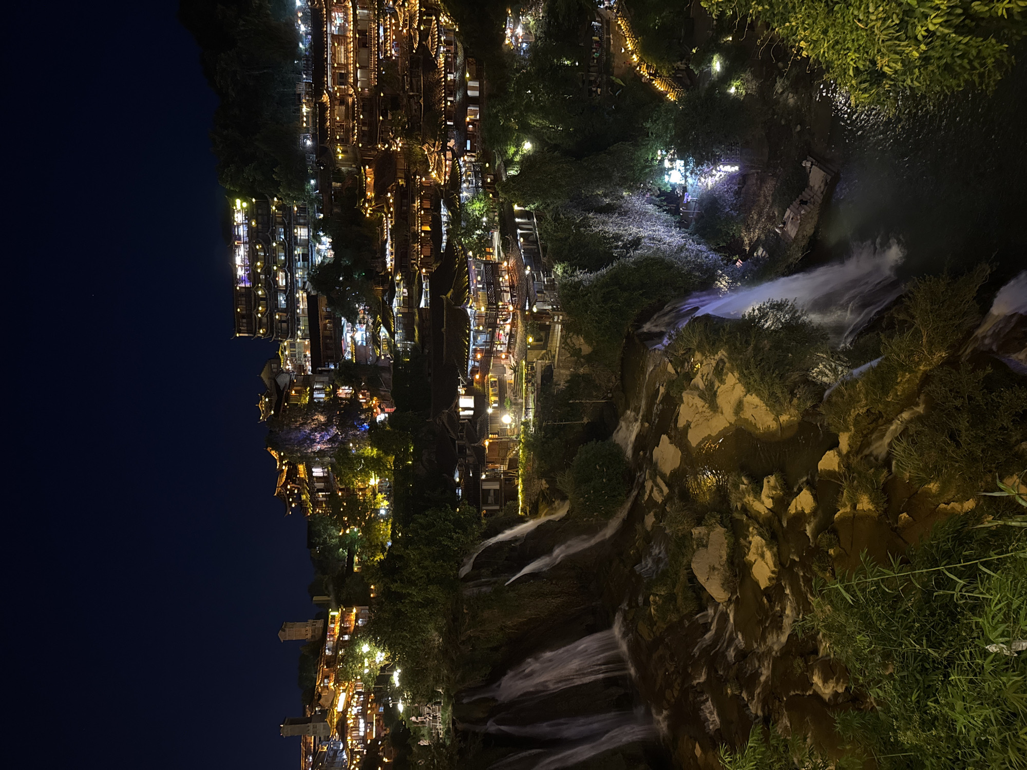 The image depicts a scenic nighttime view of the Furong Town waterfall and the illuminated ancient buildings of Furong, China, built into the cliffs above the falls. The waterfall cascades down a rocky, verdant cliff face into a dark body of water below. Several streams of water are visible, some appearing wider and more powerful than others. The water is white and frothy as it plunges.
Perched on the cliffs overlooking the waterfall are numerous traditional Chinese-style buildings, characterized by their dark wooden structures, ornate tiled roofs with upturned eaves, and glowing yellow lights. These buildings appear to be multi-storied and are built closely together, creating a dense, layered effect against the dark hillside. Some buildings have balconies and walkways, suggesting they might be hotels, restaurants, or shops. Lanterns are visible, casting a warm glow on the architecture and surrounding trees.
The foreground is dominated by the waterfall and the rocky terrain it flows over. Lush green vegetation, including trees and shrubs, is scattered throughout the scene, contrasting with the dark rock and the man-made structures. The overall impression is one of a vibrant, historic town seamlessly integrated with a dramatic natural landscape. The sky is a deep, clear dark blue, indicating it is well after sunset. There are no visible people in the immediate vicinity of the waterfall, but the lights in the buildings suggest activity within the town.