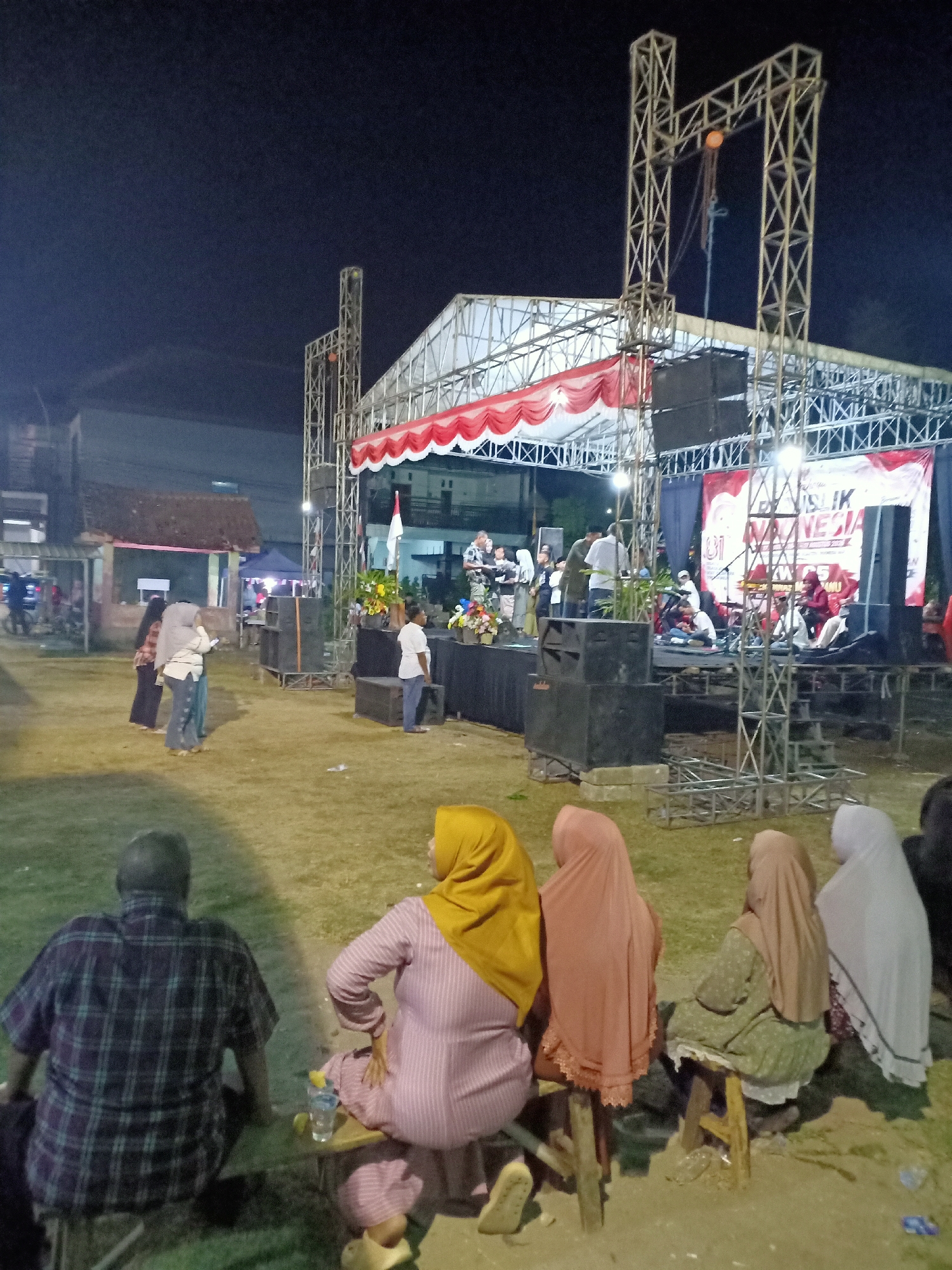 This image depicts an outdoor evening event in the city of Sumber Tlaseh, Indonesia. A stage with a red and white draped canopy and a large banner is set up, presumably for a performance. Several individuals are visible on the stage, some appearing to be musicians, while others are standing. In the foreground, a group of people are seated on benches, facing away from the camera towards the stage. The ground is covered with grass and dirt. Several speakers and lighting equipment are arranged around the stage area. The overall atmosphere suggests a community gathering or festival.