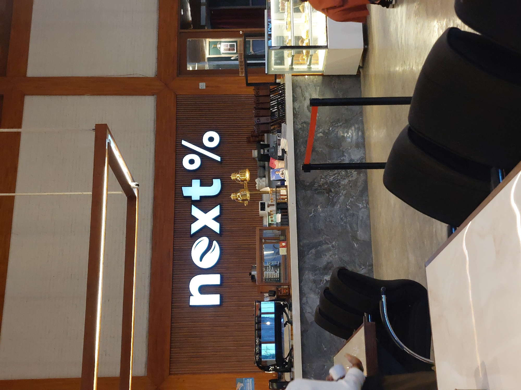 The image captures the interior of a coffee shop, likely in Pematang Siantar, Indonesia, as indicated by the presence of "next%" branding, a common local coffee chain. The scene is brightly lit, with warm wooden accents and a modern industrial feel.
Dominating the background is a large, illuminated sign spelling out "next%" in white, sans-serif font, with a stylized leaf integrated into the 'e'. Below the sign, a wooden counter displays various coffee-making equipment, including a prominent espresso machine and what appears to be a glass display case filled with pastries. The counter surface is a textured, dark gray material, possibly marble or granite.
In the foreground, several dark-colored chairs with chrome accents are visible around tables. To the left, a person, likely a customer or staff member, is partially visible, wearing white clothing and interacting with a table. The flooring appears to be a polished concrete. The overall ambiance suggests a contemporary and inviting space for enjoying coffee and refreshments.