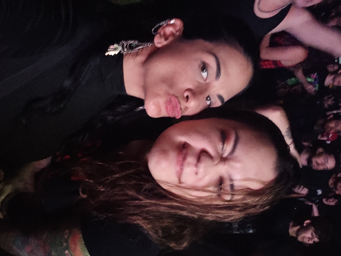 Two women are captured in a close-up at a dimly lit, crowded indoor venue, indicative of a concert or live event in Buenos Aires, Argentina. The woman on the left, with dark hair, wears a black high-neck top, a silver ear cuff, and a long, ornate silver dangle earring with white elements. She looks directly at the camera with a pouting expression. The woman on the right, with brown, wavy hair, is making a playful face, squinting her eyes and slightly pursing her lips. A colorful tattoo is visible on her left arm. The background is blurred, showing indistinct figures of a larger audience in low light conditions.