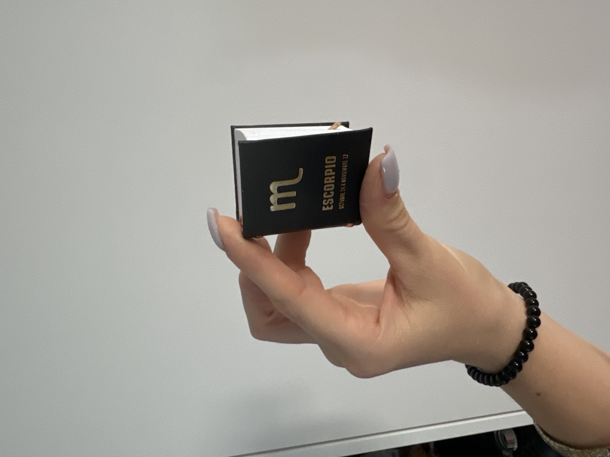 A person's hand is holding a small, black book with gold lettering. The book has the symbol for Scorpio and the text "ESCORPIO" and "OCTUBRE 24 A NOVIEMBRE 22". The hand has light purple nail polish. A black beaded bracelet is visible on the wrist. The background is a plain, light gray wall. There is no indication of the city of Murcia, Spain in the image.
