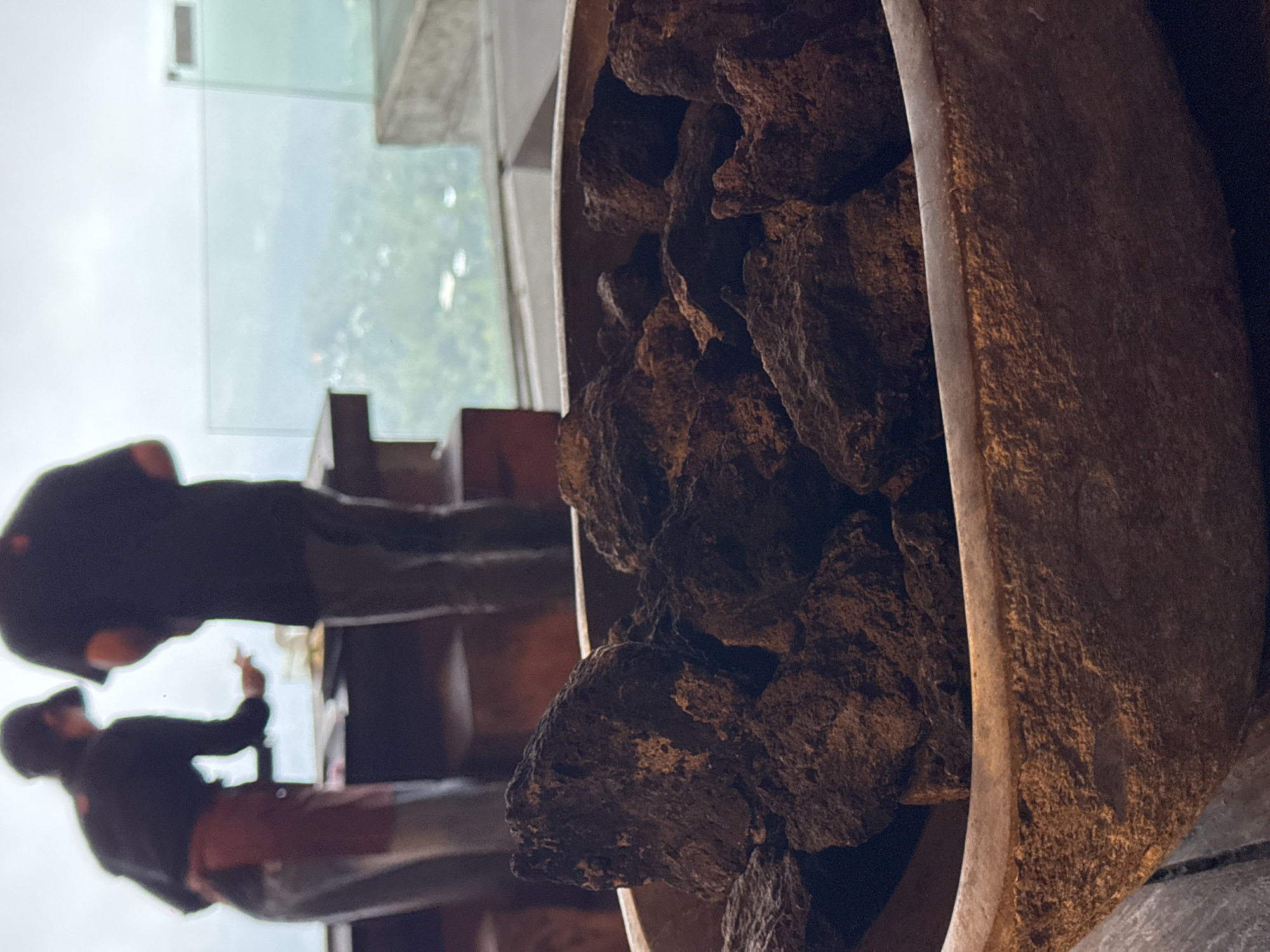 The image shows a close-up of a rustic, textured bowl filled with dark, porous rocks, likely lava rocks, placed on a wooden surface. In the background, two individuals are partially visible, standing near a counter. One person, wearing a cap and an apron, appears to be reaching towards the counter. The other person is standing beside them. Beyond them, a large window or glass barrier overlooks a lush, green landscape with some buildings visible in the distance. The overall setting suggests an outdoor or semi-outdoor dining area with a scenic view, possibly a restaurant or cafe in Kintamani, Indonesia, known for its volcanic landscapes and stunning vistas. The lighting is somewhat muted, contributing to the earthy tones of the rocks and the bowl.