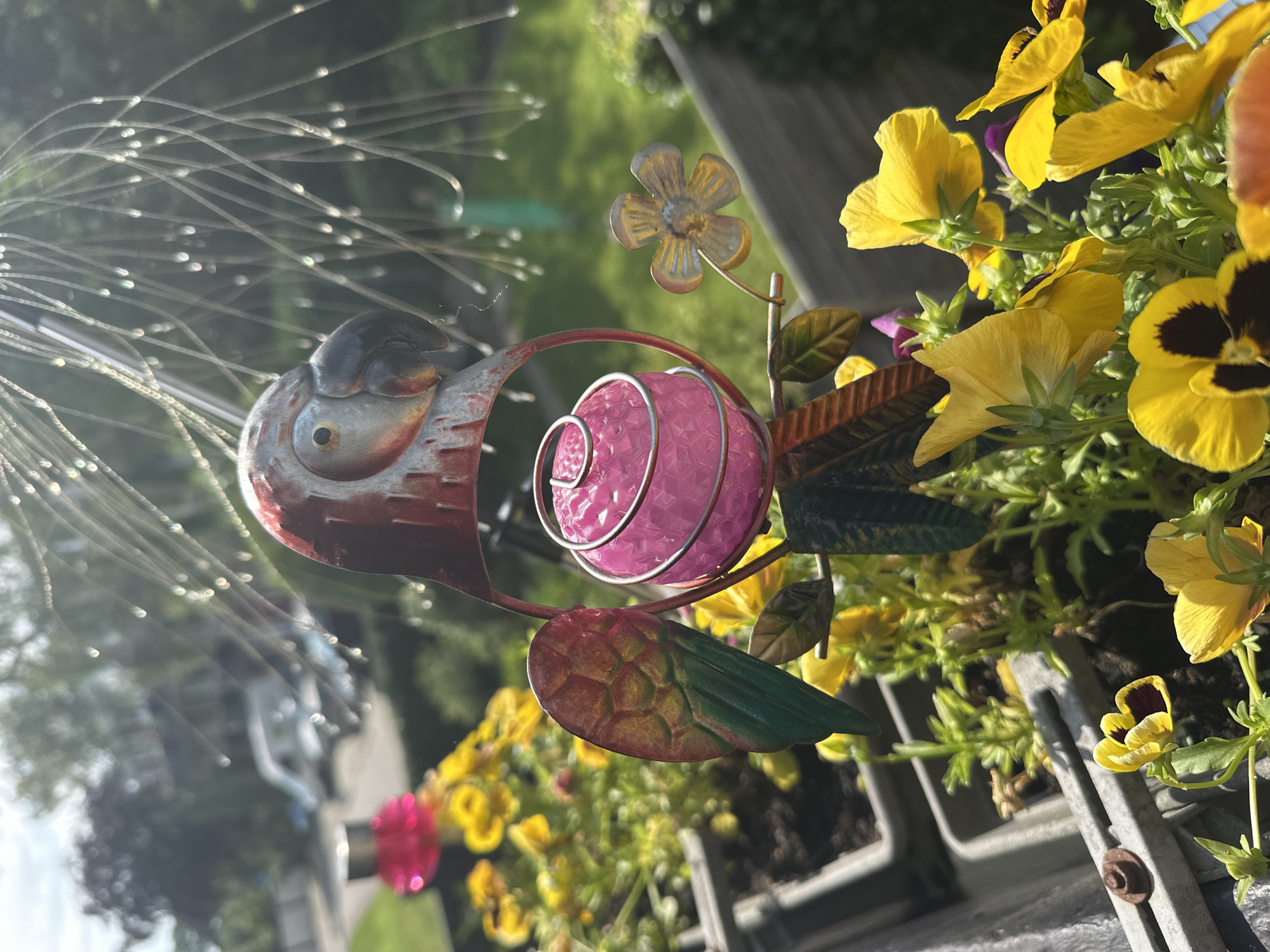 The image depicts a close-up of a metal parrot garden ornament situated amongst yellow flowers. The parrot is red and silver, with a pink orb suspended in its body. Behind the parrot, a blurry background shows green foliage and a hint of a street with a white van. The scene appears to be outdoors, likely in a garden or balcony setting. The photo was taken in Steenwijk, Netherlands.