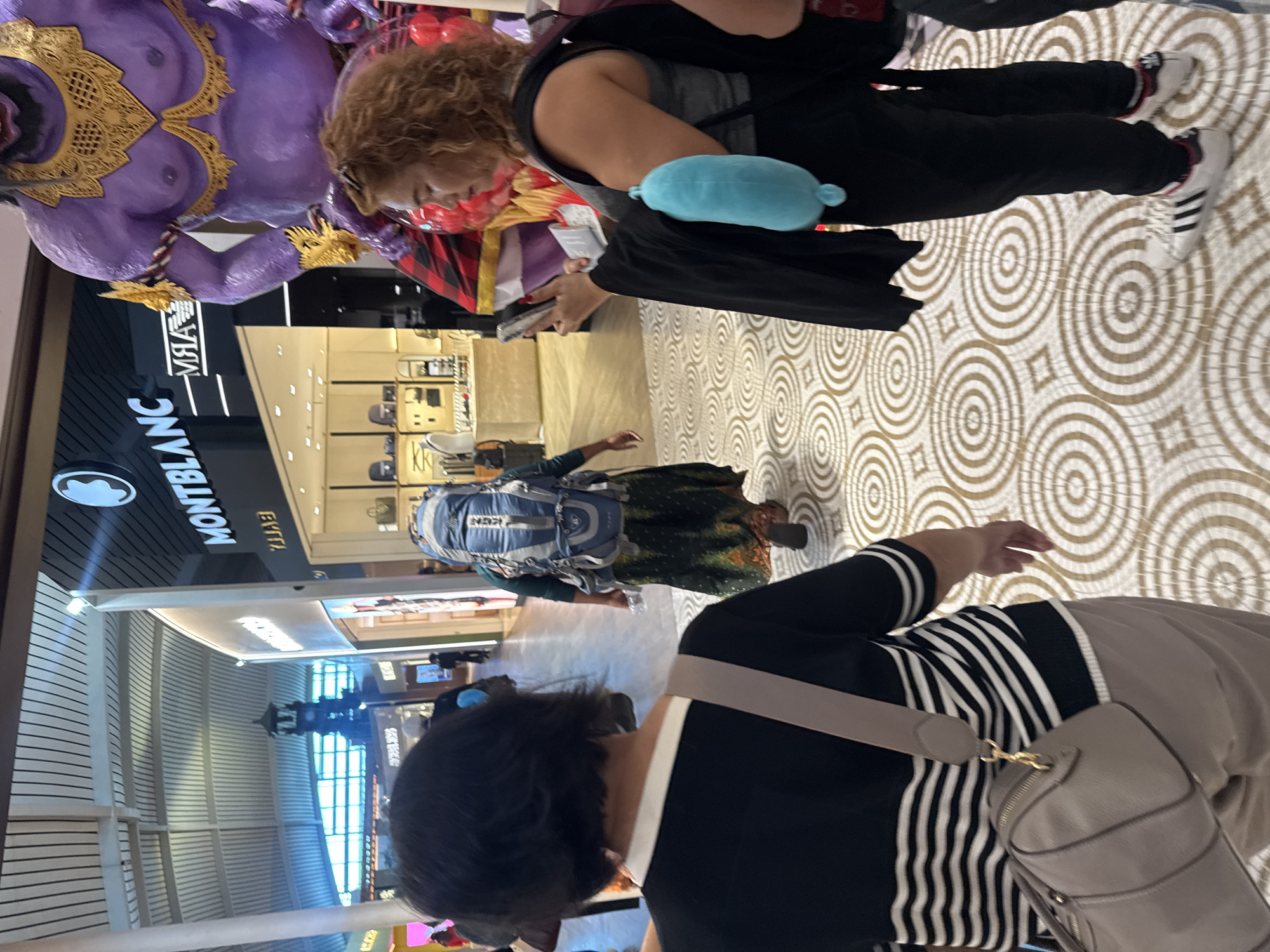 The image captures an indoor scene, likely within a shopping mall or airport terminal in Kuta, Indonesia. The distinctive floor features a mosaic pattern of light brown and off-white concentric circles.
In the foreground, the back of a person with short dark hair is visible. They are wearing a black top with prominent white vertical stripes and a beige/grey shoulder bag with gold hardware.
Moving towards the midground on the right, a person with curly light brown hair is seen from the waist up. They are wearing a grey top and black pants, holding a silver-colored mobile phone in their hand. A light blue plush toy is tucked into their waistband or bag. Their feet, wearing white Adidas sneakers with black stripes and red accents, are visible on the patterned floor.
To the left, another individual is seen from behind, carrying a large blue and grey hiking-style backpack. They appear to be wearing a green, patterned skirt or dress.
A striking element of the scene is a large, purple, statuesque figure with golden embellishments, depicting a bare-chested deity or mythical creature, located on the far left.
In the background, a storefront sign prominently displays "MONTBLANC" in white letters against a black background. Below it, the word "EYIYA" is visible but appears reversed, suggesting it might be reflected or a stylized part of the signage.
The overall atmosphere suggests a busy public space, with people moving through, possibly engaging in shopping or travel activities. The lighting is bright and even, indicative of an interior setting during the day.