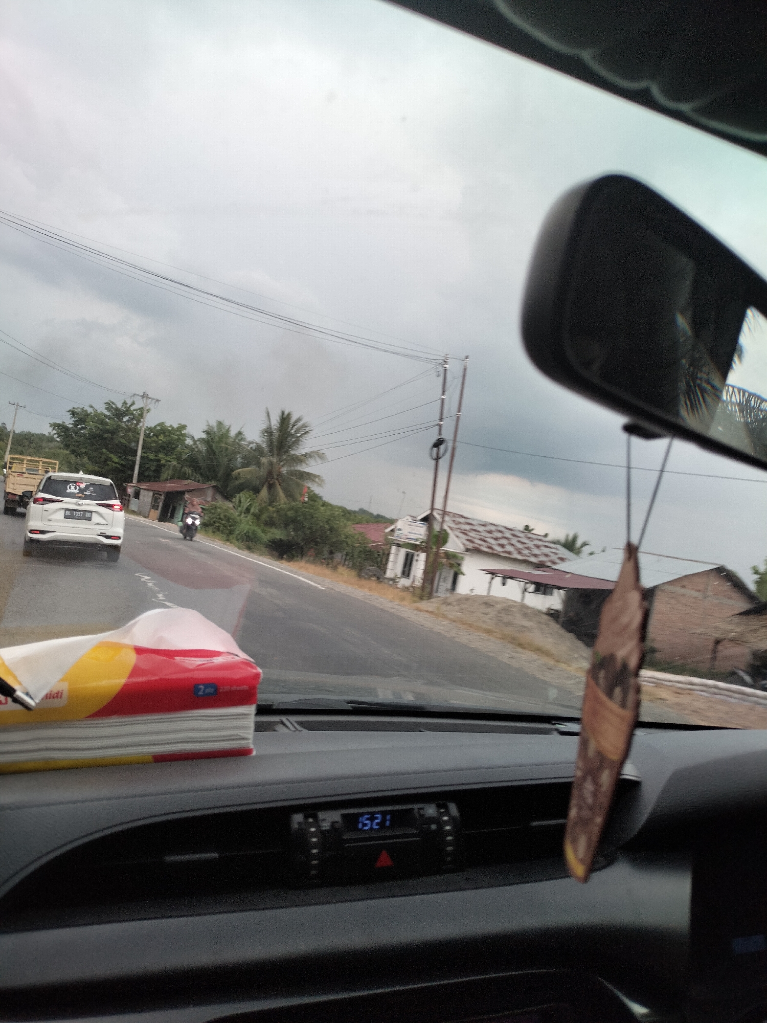 An overcast sky looms above a two-lane road in Bukit Selamat, Indonesia. The perspective is from inside a vehicle, looking forward through the windshield. In the foreground, on the dashboard, sits a box of tissues with red, yellow, and white packaging. To the right, a car's rearview mirror hangs from the ceiling, and a brown, leaf-shaped air freshener dangles from it.
The road itself is asphalt, with a yellow line marking the center. A white car with license plate DL 1357 DRG is driving ahead in the left lane, with a truck behind it. A motorcycle rider is visible in the right lane, further down the road. On the right side of the road, several houses with corrugated metal roofs and brick and wooden walls are scattered amongst lush green vegetation and palm trees. Electrical poles and wires run parallel to the road. The overall scene suggests a rural or semi-rural environment under a cloudy sky.
