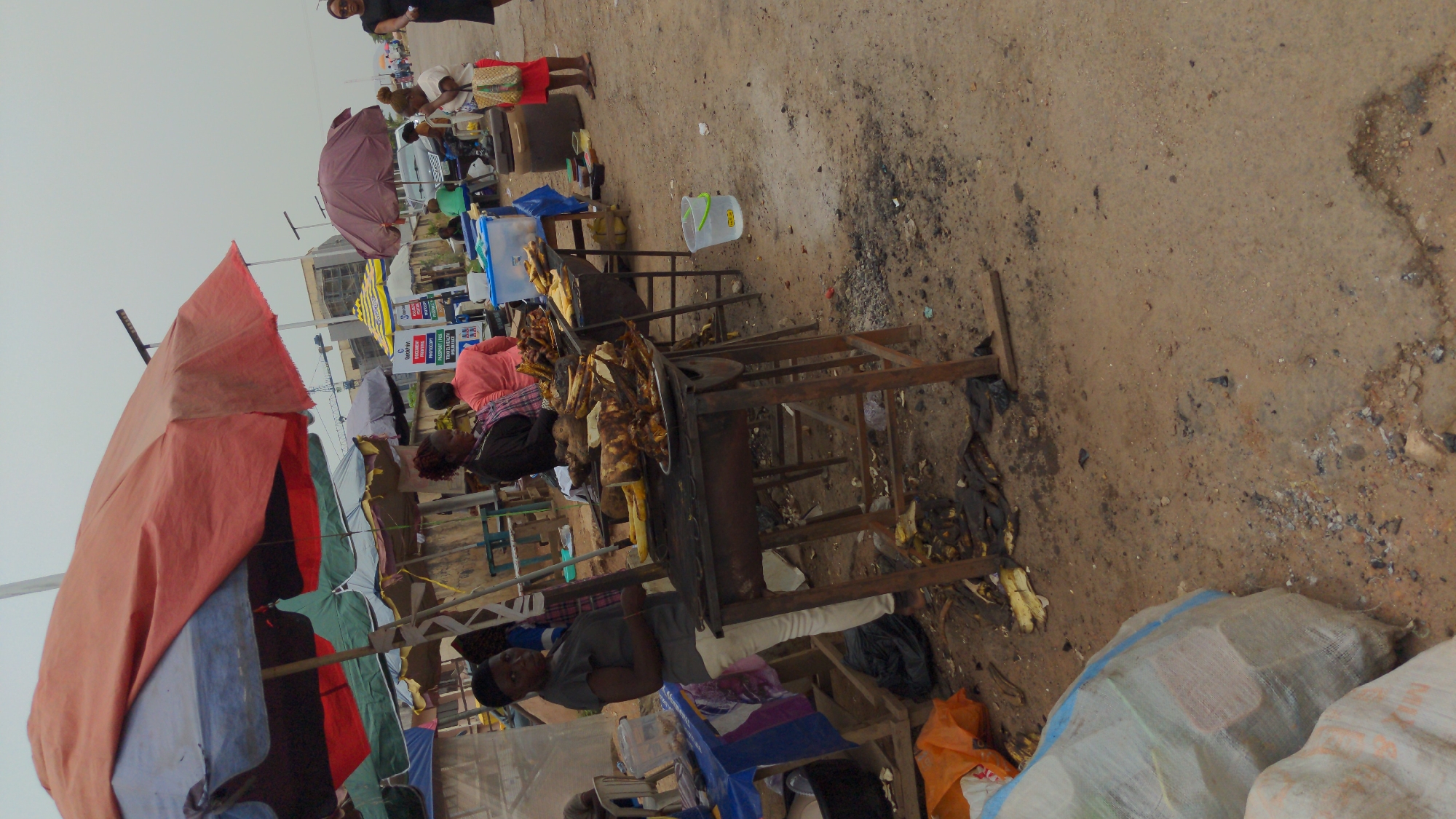 This image captures a bustling outdoor market or street vendor scene, likely in Abuja, Nigeria. The perspective is slightly elevated, looking down across an area with multiple makeshift stalls on an uneven dirt ground.
Several people are visible engaging in daily market activities. In the mid-ground, a person wearing a grey top sits behind a stall, appearing to attend to customers or goods. Further back, another person in a pink top is also stationed behind a stall. Towards the background, a woman in a black dress walks away, while another woman in a white top and red skirt stands near a stall.
The market is characterized by numerous stalls, many constructed with wooden frames. Large, colorful umbrellas, including a prominent red/orange one in the foreground and others in blue, green, purple, and grey, provide shade. A central focus is on food vending; one stall displays what appears to be roasted plantains or yams, and possibly grilled meat or fish, indicating a street food operation. Various plastic containers, buckets (one labeled "FOODGRADE" with a green handle), and mesh bags containing goods are scattered around and on the stalls.
The ground is unpaved, covered in dirt and sand, with visible debris such as ash, charred remnants from cooking, and discarded organic matter like banana peels. The overall atmosphere suggests a functional, everyday market environment. The sky is overcast or hazy, indicating daytime but not necessarily bright sunlight.
In the background, a blue and white sign is partially visible with text including "TYPHOON," "INVESTMENT," "PROPERTY," "PROGRAMME," "TRAVEL AGENCY," "TOURISM," and "HOTELS," suggesting that formal businesses or services may be located nearby or advertised within the market area.