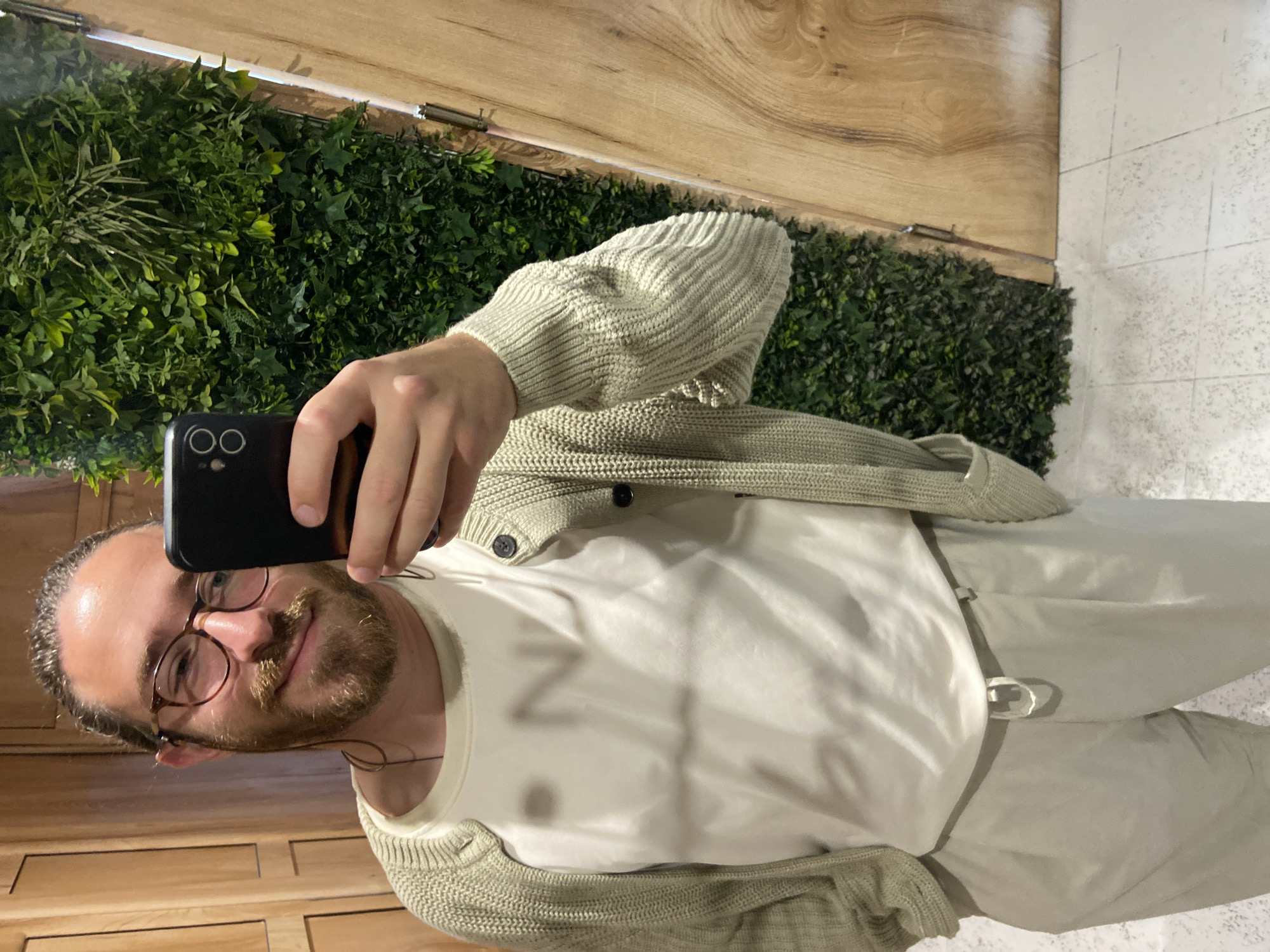 A man with fair skin, a beard, and glasses is taking a selfie in what appears to be a bathroom. He's wearing a white t-shirt, a light-colored cardigan, and light-colored pants with a belt. The background consists of a wall covered in greenery, a wooden paneled wall, and a tiled floor. There is no information in the image to confirm this is in Petrovac centar, Montenegro.