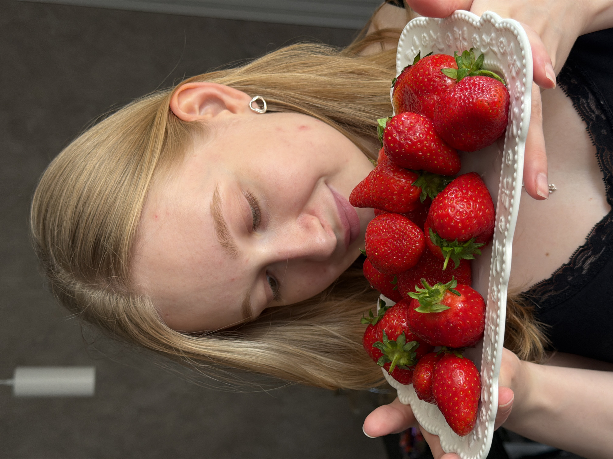 A young woman with blonde hair is depicted holding a white, ornate serving plate filled with approximately 15-20 ripe, red strawberries. Her head is tilted downwards, and her gaze is directed towards the fruit, exhibiting a subtle smile. She wears a black garment featuring a lace trim along the neckline, a small silver-colored pendant on a thin chain, and a silver-colored, open-ended, irregular heart-shaped earring in her left ear. Her hands support the white plate, which is adorned with raised decorative patterns and a scalloped edge. The background is an indistinct indoor setting, characterized by a dark, plain wall on the left and a lighter-colored vertical surface on the right. A white cylindrical object, consistent with a suspended light fixture, is visible in the upper left portion of the blurred background. The image contains no discernible geopolitical markers to confirm the specific location as Rossiyskiy, Russia.