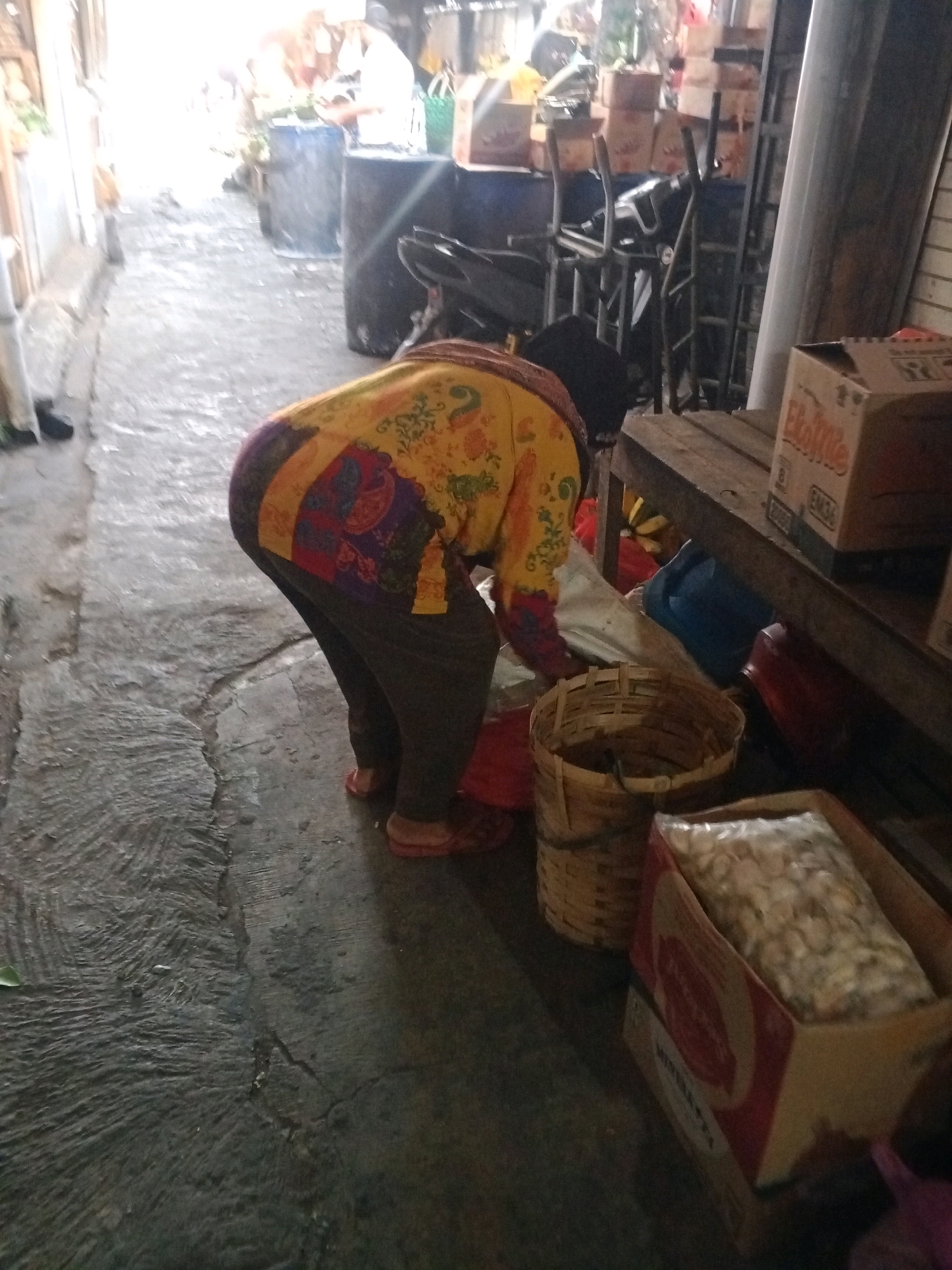 A person, wearing a colorful patterned shirt and dark pants, bends over a woven basket on a paved ground. Behind the person, various items are visible, including a motorcycle, stacked boxes, and a wooden table. The environment appears to be an outdoor market or storage area, possibly in Wonogiri, Indonesia, given the context. The lighting is dim, suggesting it might be an overcast day or an indoor/covered space.