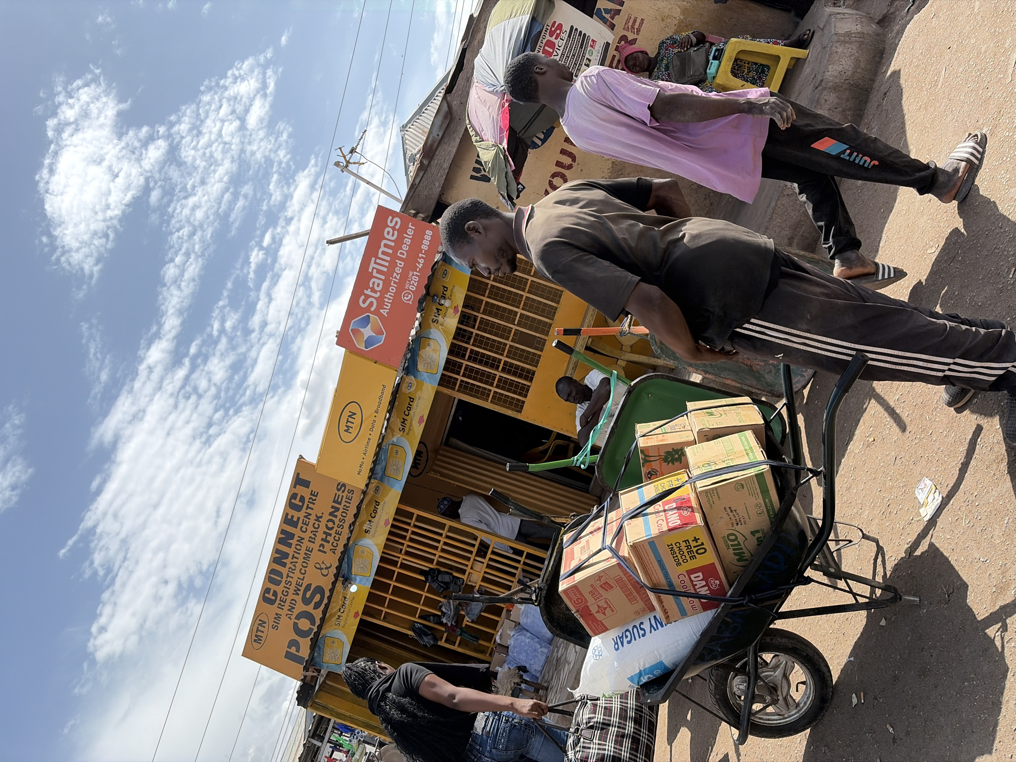 The image captures a vibrant outdoor street scene in what appears to be a commercial area, likely in Jos, Nigeria, under a bright, sunny sky with scattered white clouds. The ground is unpaved, suggesting a market or informal trading zone.
In the foreground, a wheelbarrow is heavily loaded with various goods, including cardboard boxes labeled "DANO," "MILO," and "Nestlé," along with a white sack clearly marked "SUGAR." A woman with long dark braids is positioned as if guiding the wheelbarrow, while a man in a dark brown short-sleeved shirt leans over it, seemingly adjusting the load. Another man, wearing a light pink shirt, black track pants with "JUNT" visible, and striped sandals, stands nearby, looking towards the right.
The background features several modest buildings housing shops or service centers. Prominent yellow and red signs are visible. A large yellow sign reads: "MTN CONNECT SIM REGISTRATION CENTRE AND WELCOME BACK. POS & PHONES ACCESSORIES." Smaller MTN signs advertise "MoMo Airtime Data Broadband" and "SIM Card Registration Center." A red "StarTimes Authorized Dealer" sign displays a phone number: "0201-461-8888." Another sign lists "NEPOINT SERVICES" including "CASH DEPOSIT," "CASH WITHDRAWAL," "GO TV," "BUY AIRTIME," and "MONEY TRANSFER."
In the distance, other individuals are visible; one person is seen resting or sleeping on a yellow plastic chair. Utility poles and overhead power lines crisscross the sky. The bright daylight creates strong shadows on the ground. The scene depicts everyday commercial activity and transport in an urban African setting.