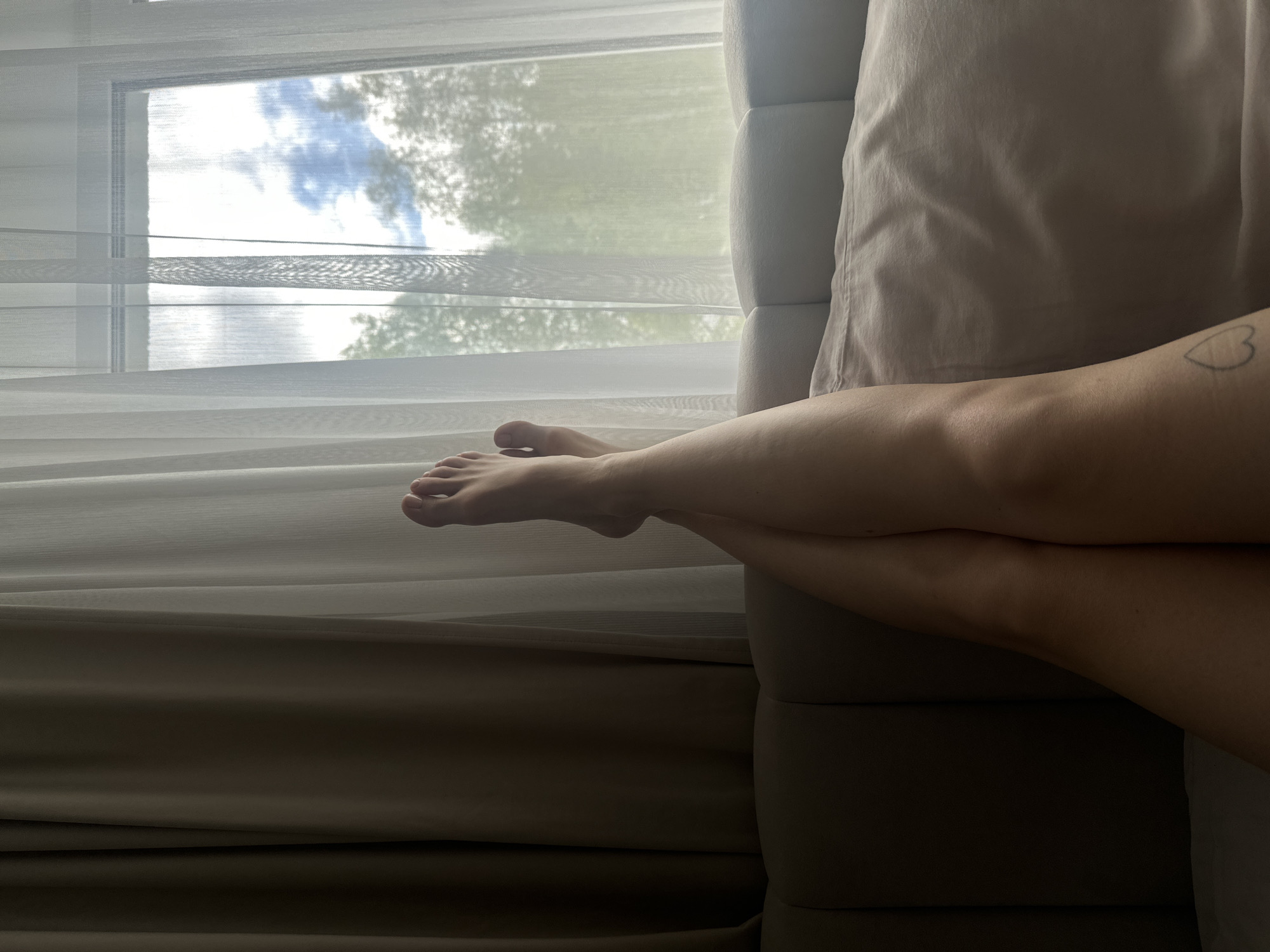 A pair of crossed legs rests on a beige sofa. A heart-shaped tattoo is visible on the right leg. The legs extend toward a window covered by sheer curtains. Outside the window, trees and a cloudy sky are visible. The location cannot be determined based on the image.