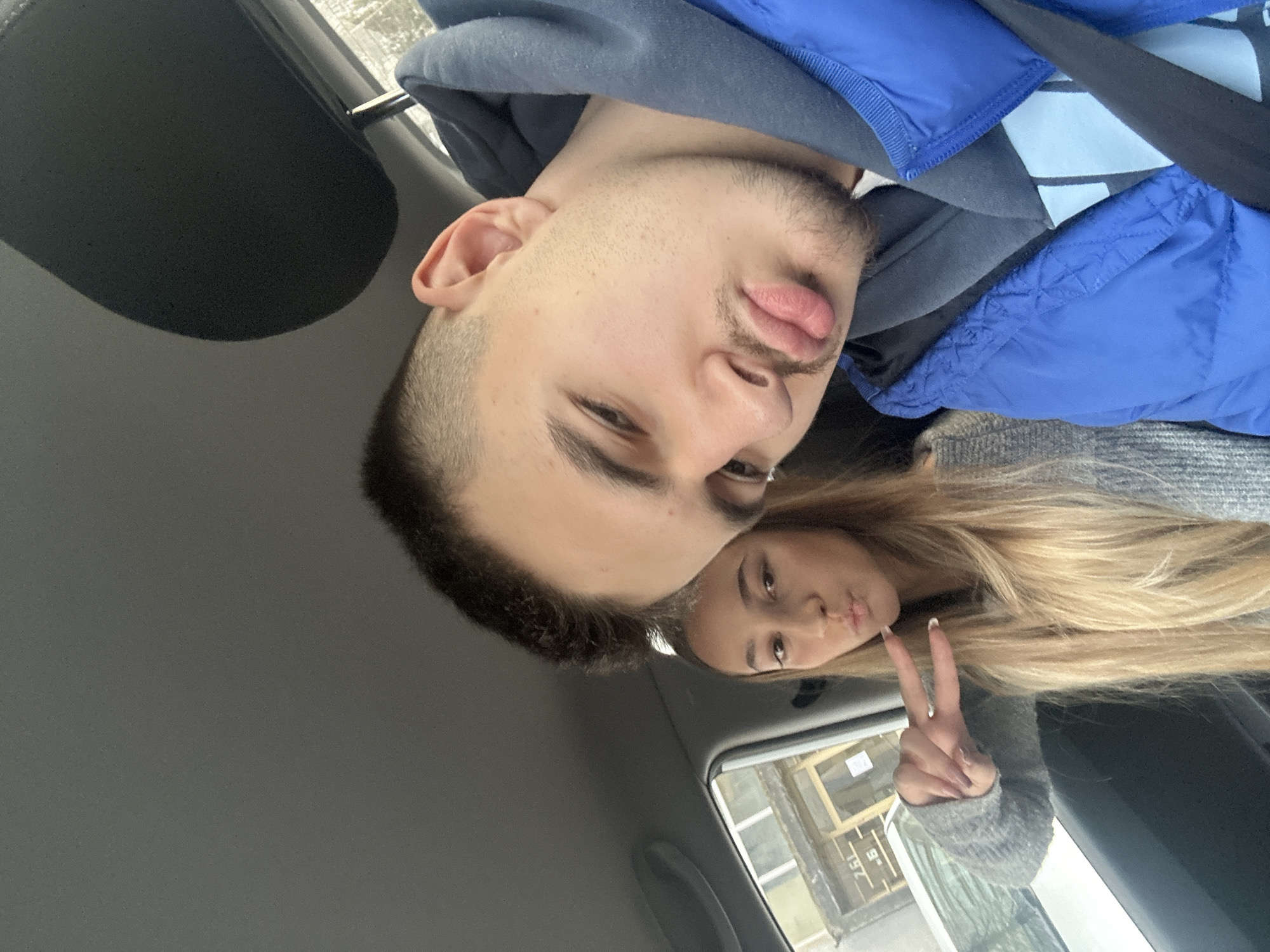 Two individuals are in a car. The man has short, dark hair, a beard, and is wearing a blue hoodie. He is sticking his tongue out slightly. A blonde woman is behind him, holding up a peace sign. The location is a city, potentially Sofia, Bulgaria, but this cannot be determined from the image alone. The interior of the car is visible. A window shows a building outside.