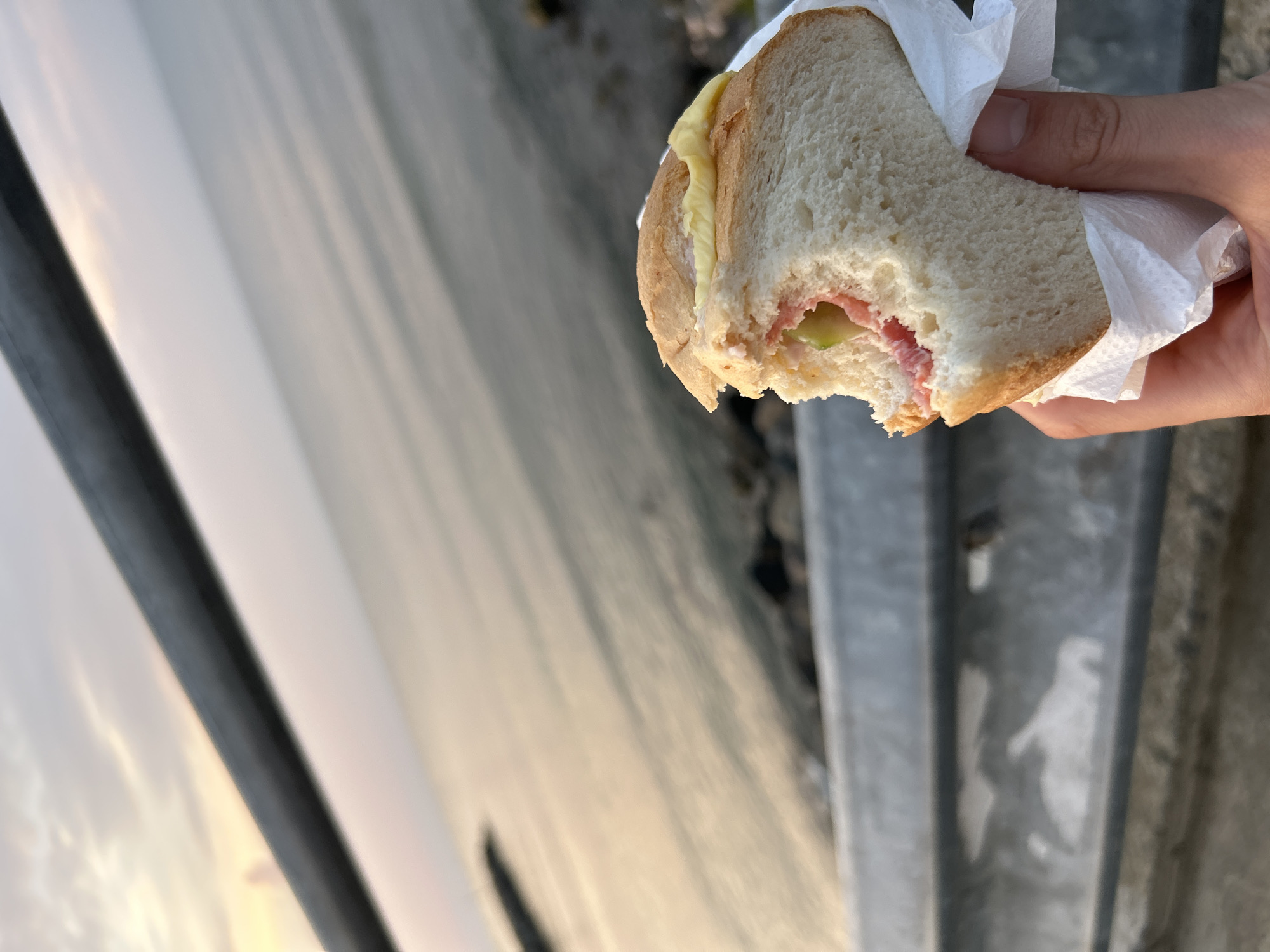 A hand holds a partially eaten sandwich wrapped in white paper, with a bite taken out of it. The sandwich appears to contain pink ham and green slices. The background is a blurry body of water and sky, possibly at sunset, with a metal railing in the foreground. The scene is set in Ravda, Bulgaria.