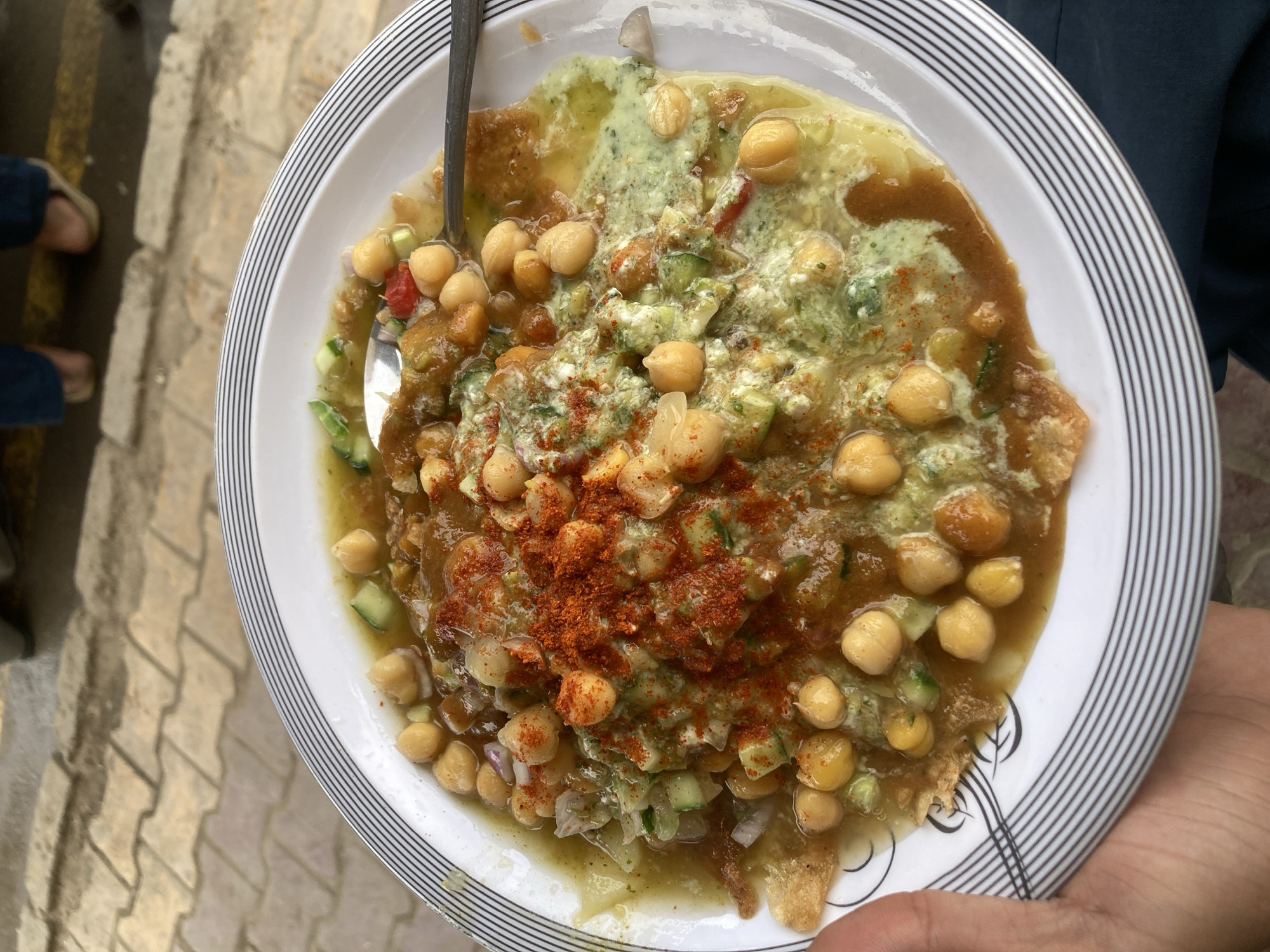 A plate of chana chaat, a popular South Asian street food, is held by a person. The chaat consists of chickpeas, chopped vegetables (like cucumber and onion), sauces (brown and green), and a sprinkle of red chili powder, all arranged on a fried base. A spoon is visible in the dish. The plate has a white background with a black-striped rim. The photograph appears to have been taken outdoors, possibly in Quetta City Tehsil, Pakistan, based on the prevalence of this dish in the region. A person's feet can be seen in the periphery with an adjacent sidewalk. The person holding the plate is wearing a dark-colored shirt.