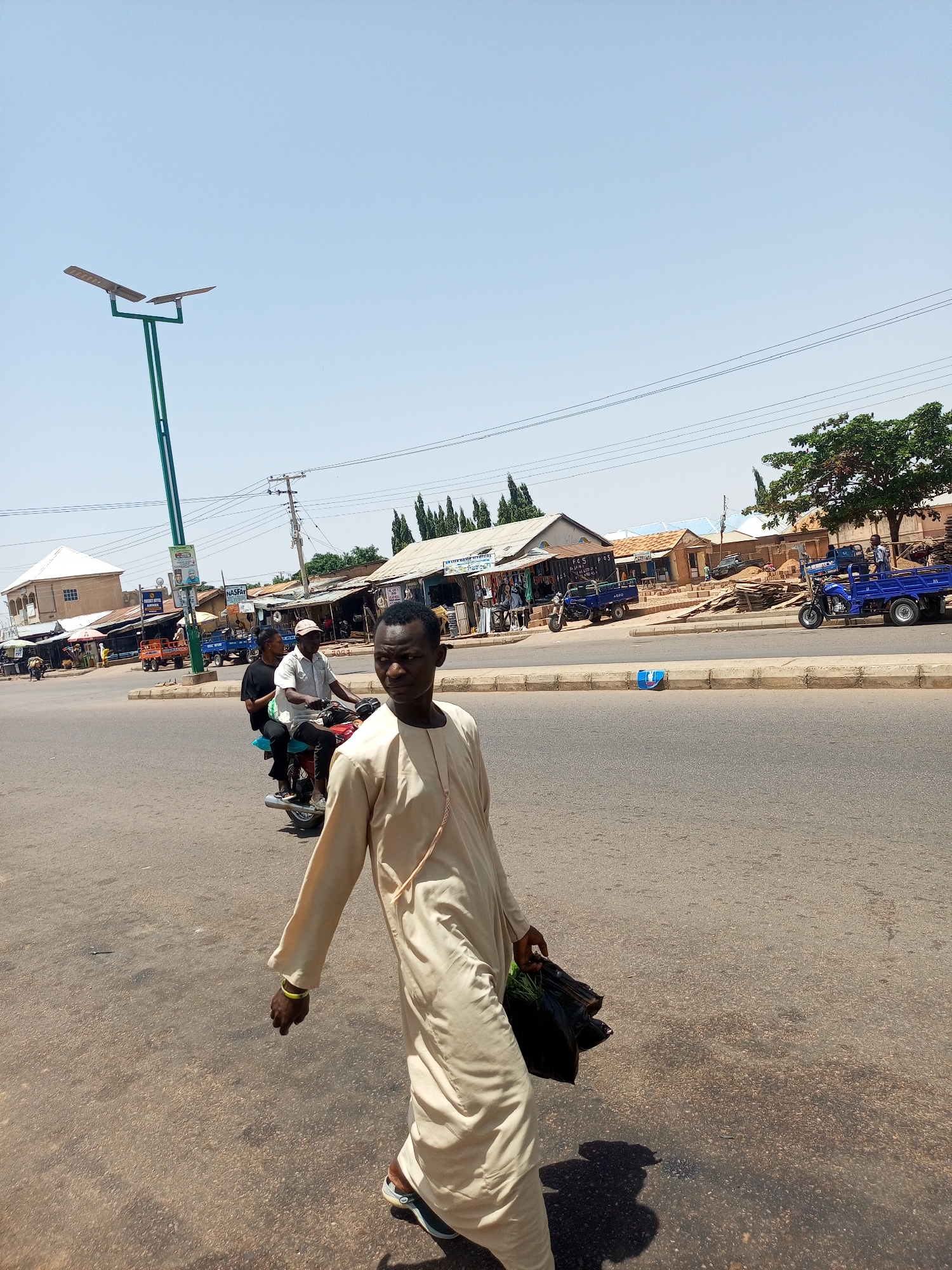 This media file depicts a daytime scene on a paved road in Tumfure, Nigeria. The weather appears to be sunny and clear. The primary subject in the foreground is a man walking towards the right of the frame. He is wearing a light-colored traditional robe and carrying a black plastic bag filled with green produce in his left hand. His right hand has a yellow bracelet.
In the background, several other elements contribute to the scene. A motorcycle carrying two passengers is moving from left to right, behind the man in the foreground. Further in the background, there are various buildings that appear to be shops or market stalls lining the road. Some of these structures have corrugated metal roofs and open fronts displaying goods. A blue truck with a flatbed is parked on the right side of the road, and another blue truck is visible further down the road. A lamppost with two solar panels stands prominently on the left side of the image. Power lines are also visible stretching across the sky. The general impression is a bustling roadside market environment.
Visible text includes "NASFAT" on a sign in the background. The overall mood of the image is neutral, capturing a slice of everyday life in this location. The time of day is clearly indicated by the bright sunlight and the long shadows cast.