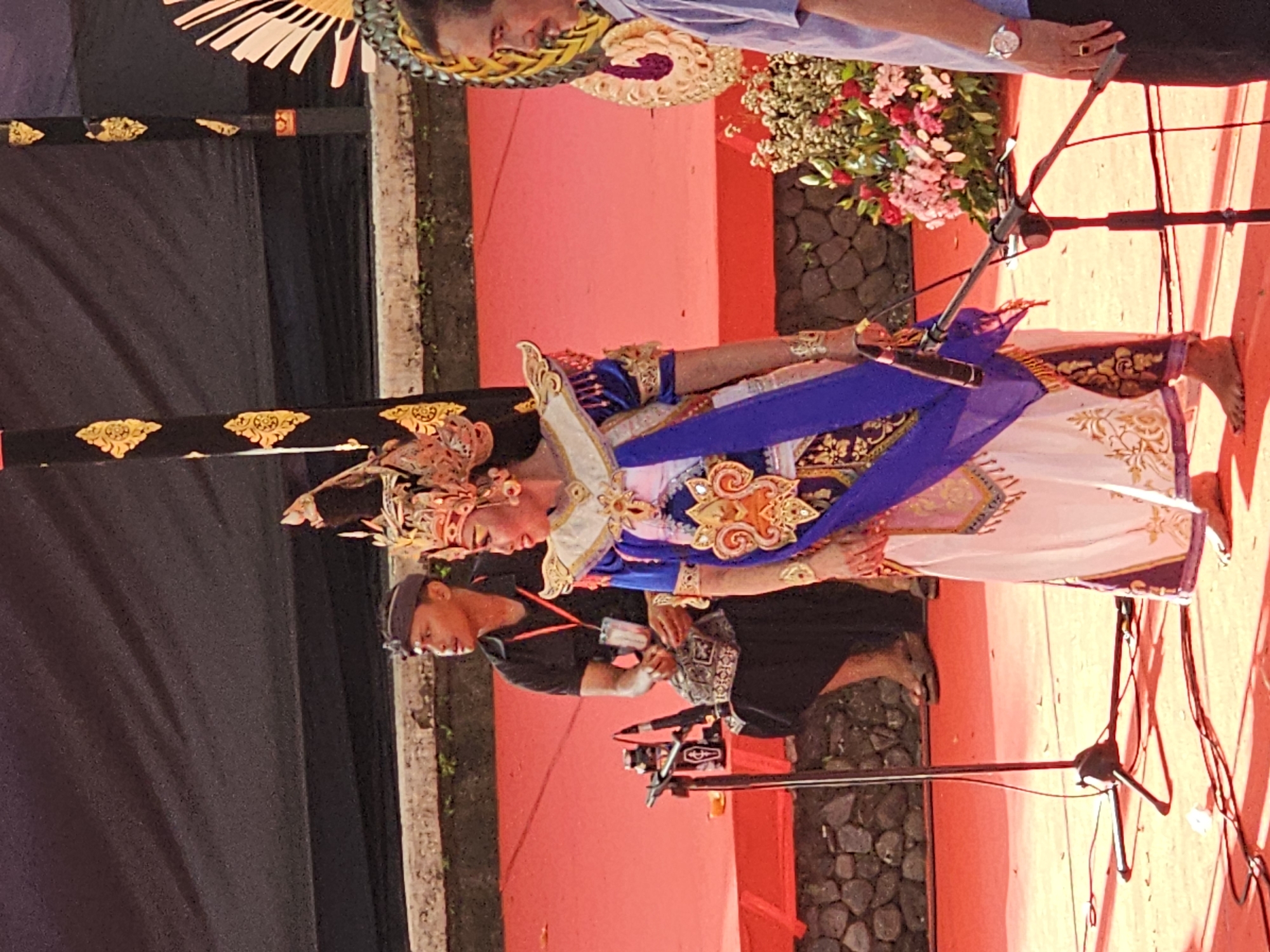 A full shot captures a Balinese dancer in elaborate costume and headdress standing on a red carpeted stage, positioned in front of a dark draped backdrop. The dancer, facing right, has elaborate gold and blue embroidery on her traditional attire. A microphone stand is in the foreground. To the left of the dancer, a man in a black t-shirt and head covering, identified as a security personnel, is seated. In the background, a cluster of pink and white flowers is visible. The scene is indicative of a cultural performance, possibly a traditional dance or ceremony. The location in Denpasar, Indonesia, suggests a cultural event.
