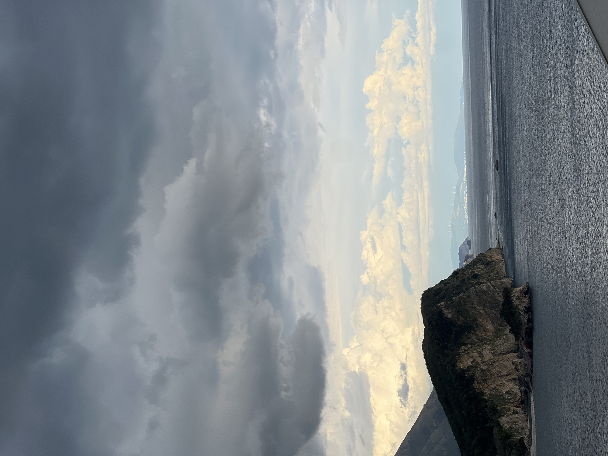 This media file captures a dramatic coastal landscape, likely viewed from the sea or a high vantage point overlooking the water, consistent with the location context of Budva, Montenegro.
In the foreground and mid-ground, a vast expanse of dark, rippling sea dominates the lower and right portions of the frame. On the right side, a prominent, rugged rocky island or headland rises steeply from the water. Its dark, textured rock faces show various strata and are sparsely covered with vegetation.
The upper and left parts of the image are filled with a dynamic sky. On the far left, heavy, dark grey storm clouds create a sense of impending weather. These gradually transition towards the middle and right into brighter, more illuminated cumulus clouds, catching a warm, possibly golden light. This contrast suggests either a late afternoon/early morning sun breaking through the clouds or a rapidly changing weather pattern.
Along the distant horizon, a faint, low-lying coastline is visible, featuring small, indistinct structures that could be buildings or wind turbines. The overall impression is one of natural grandeur and a slightly moody atmosphere due to the contrasting sky elements. There is no visible text or human activity depicted.