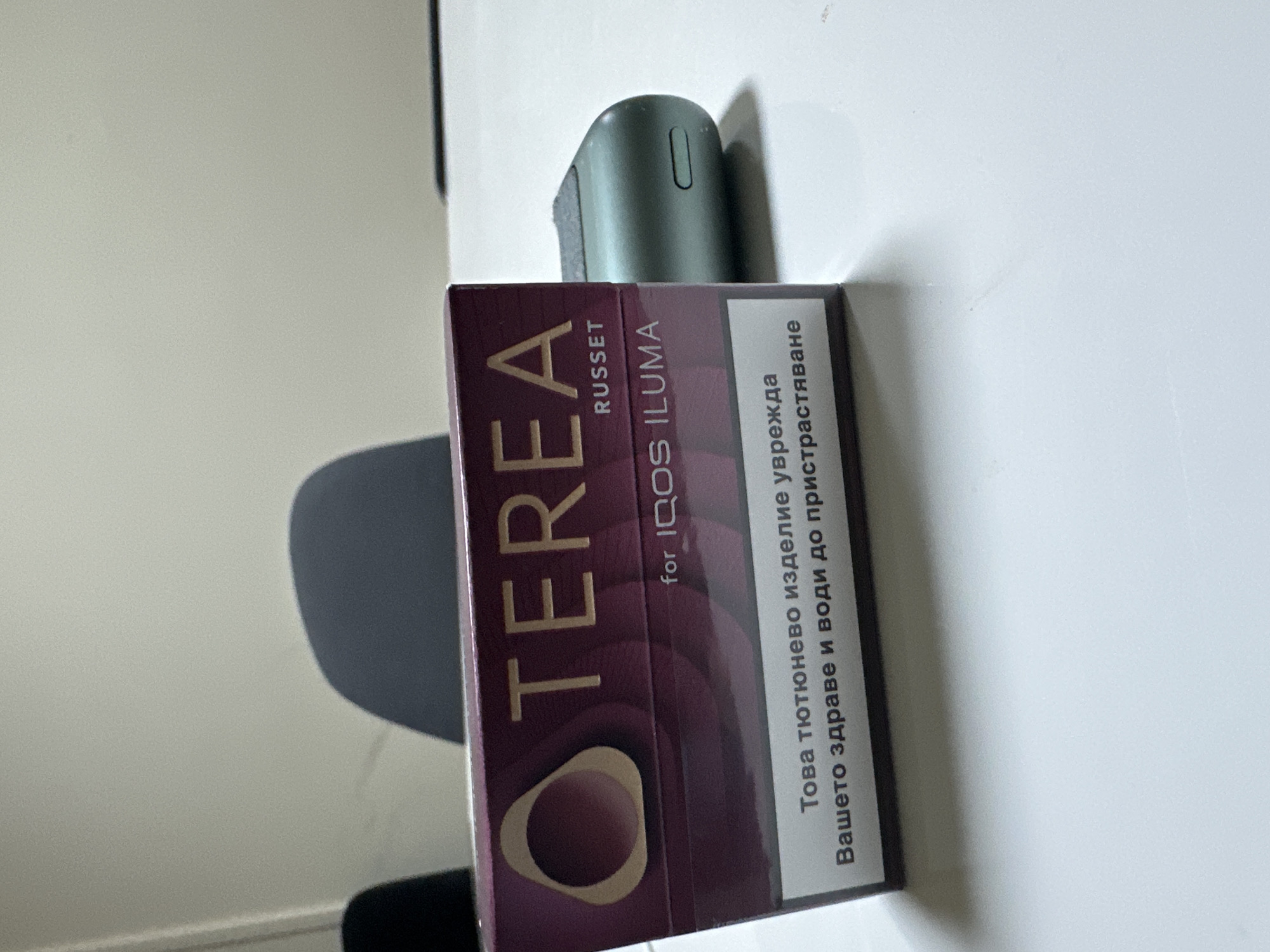 The image captures a still-life arrangement featuring a box of "TEREA" tobacco sticks and a part of an IQOS ILUMA device. The box, predominantly burgundy with gold accents, is positioned horizontally in the foreground. "TEREA" is printed prominently in large capital letters, with "RUSSET" and "for IQOS ILUMA" also visible.
Resting partially on top of the TEREA box and behind it is a sleek, green or teal-colored IQOS ILUMA device, showing part of its top and a charging or indicator slot. The background consists of a plain, light-colored wall, possibly beige or light grey, with a distinct shadow from what appears to be a chair or a similar object cast upon it. The items are placed on a flat, light-colored surface, likely a table or desk.
A crucial detail is the health warning printed in Bulgarian on the side of the TEREA box. The text reads: "Това тютюнево изделие уврежда Вашето здраве и води до пристрастяване." This translates to "This tobacco product harms your health and leads to addiction," consistent with the given location context of Sofia, Bulgaria. The lighting is even, suggesting an indoor setting with either natural diffused light or artificial light, with no specific time of day discernible. No people are present, and no particular activity is taking place beyond the display of the products.