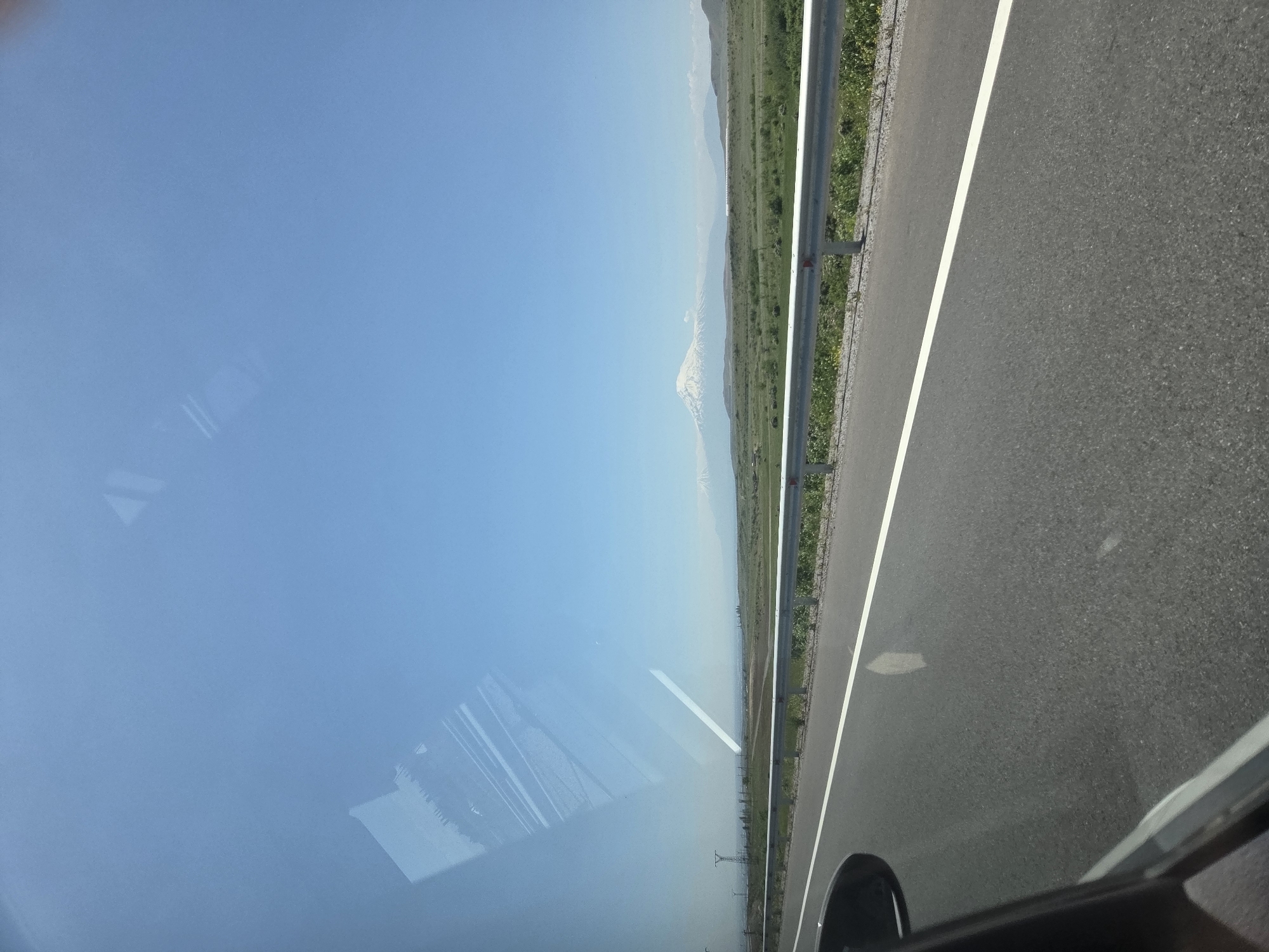The image presents a bright, clear daytime view captured from inside a vehicle, looking out across a rural or semi-rural landscape. The scene is dominated by a vast blue sky in the upper portion. In the middle ground, green fields and rolling terrain extend towards the horizon. In the distance, a majestic snow-capped mountain, likely Mount Ararat given the Kosh, Armenia context, is visible, its peak appearing white against the lighter blue, hazy distant sky.
A grey metal guardrail runs along the right side of the frame, separating the green roadside from the paved asphalt road. The road features a distinct white solid line. In the bottom right corner, a portion of the vehicle's dark interior, possibly a dashboard or door frame, and a wheel arch are visible. The left side of the image is affected by significant reflections on the vehicle's window, showing abstract light-colored shapes, including parallel lines and angular forms, obscuring parts of the exterior view. The weather appears sunny and clear. There is no visible text in the image.