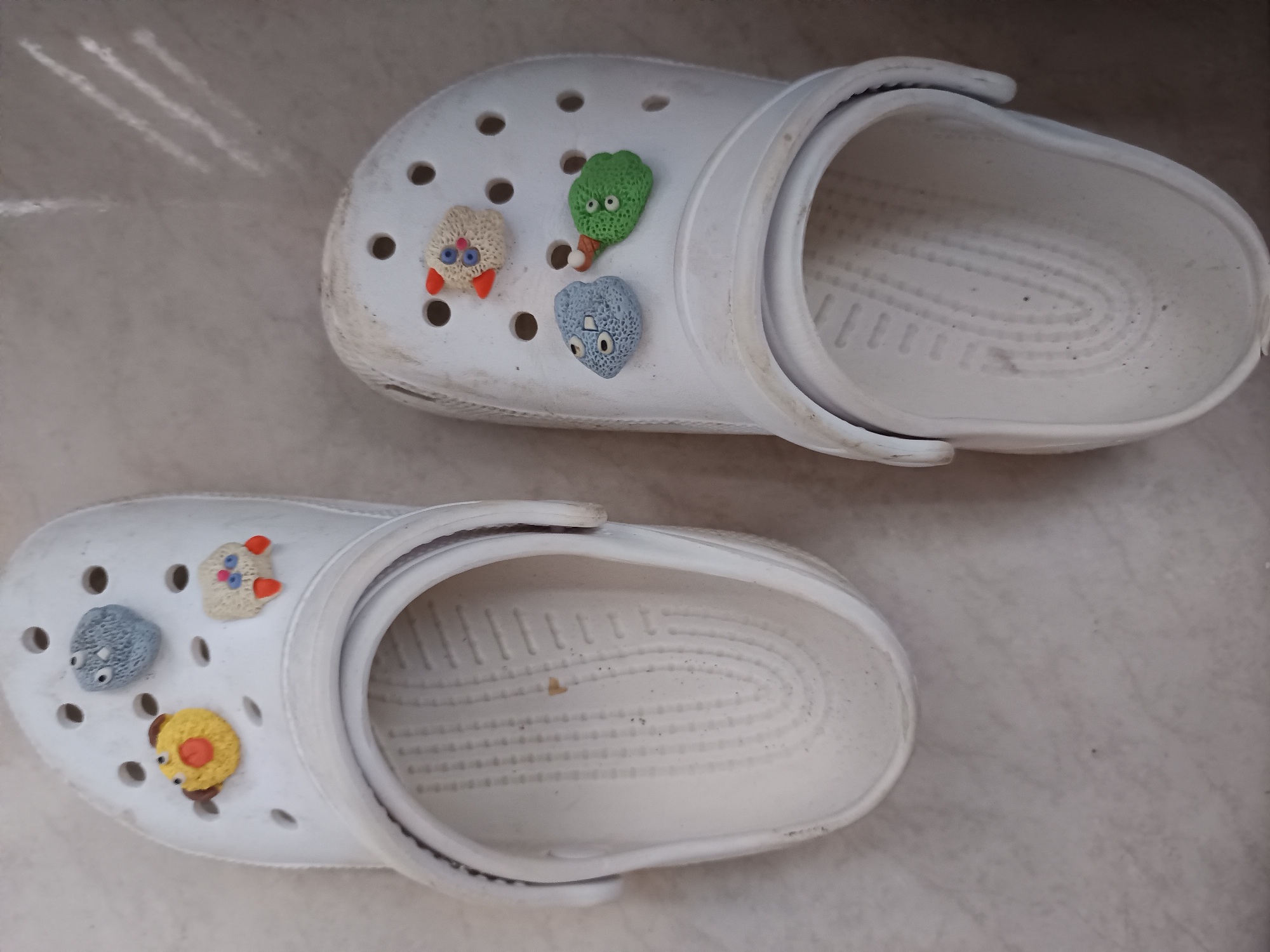 The image shows two white Crocs shoes lying on a tiled surface. Each shoe has several cartoonish charms attached to the top. The charms appear to be small, colorful, and feature simple designs. The shoes are slightly dirty and show signs of wear. The location is unknown but has been designated as Maleber, Indonesia.
