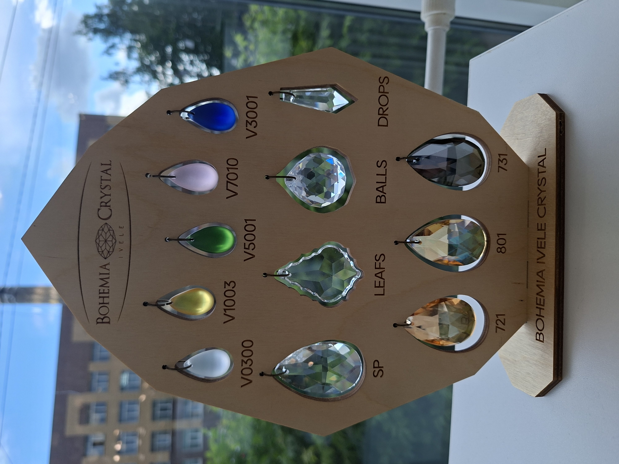This is a close-up, eye-level, exterior photograph taken on a bright, sunny day. The image depicts a wooden display stand featuring various crystal pendants. The stand is hexagonal in shape and made of light-colored wood, with the "BOHEMIA CRYSTAL IVELE" logo engraved at the top.
There are three rows of crystal pendants. The top row displays five small, colored, teardrop-shaped pendants in white, yellow, green, pink, and blue. Each pendant has a corresponding label below it: V0300, V1003, V5001, V7010, and V3001.
The middle row features four larger, faceted crystal pendants, with a distinct teardrop shape, a unique irregular shape, a round faceted shape, and a thin, elongated shape. These are labeled SP, LEAFS, BALLS, and DROPS, respectively.
The bottom row consists of three teardrop-shaped pendants in shades of amber and dark gray, labeled 721, 801, and 731.
The display stand is placed on a white surface, and in the background, through a window, a cityscape is visible. This includes a multi-story brick building with numerous windows and a tall, industrial chimney or structure. Trees and a blue sky with some clouds are also part of the background. The overall scene suggests a retail or exhibition setting, likely a showroom or a market stall, where these crystal items are being presented for sale or display. The photograph was taken in the city of Izhevsk, Russia.