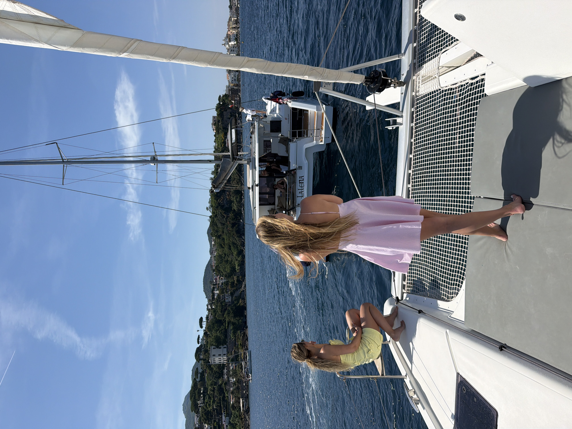 Two fair-skinned young women are on a catamaran on the water near Ischia, Italy. One woman wears a pink and white striped dress; the other wears a yellow dress. A similar catamaran, with the name "Lalena" on its side, is nearby. The water is choppy, and the shore is lined with buildings and trees. The sky is blue with light clouds.