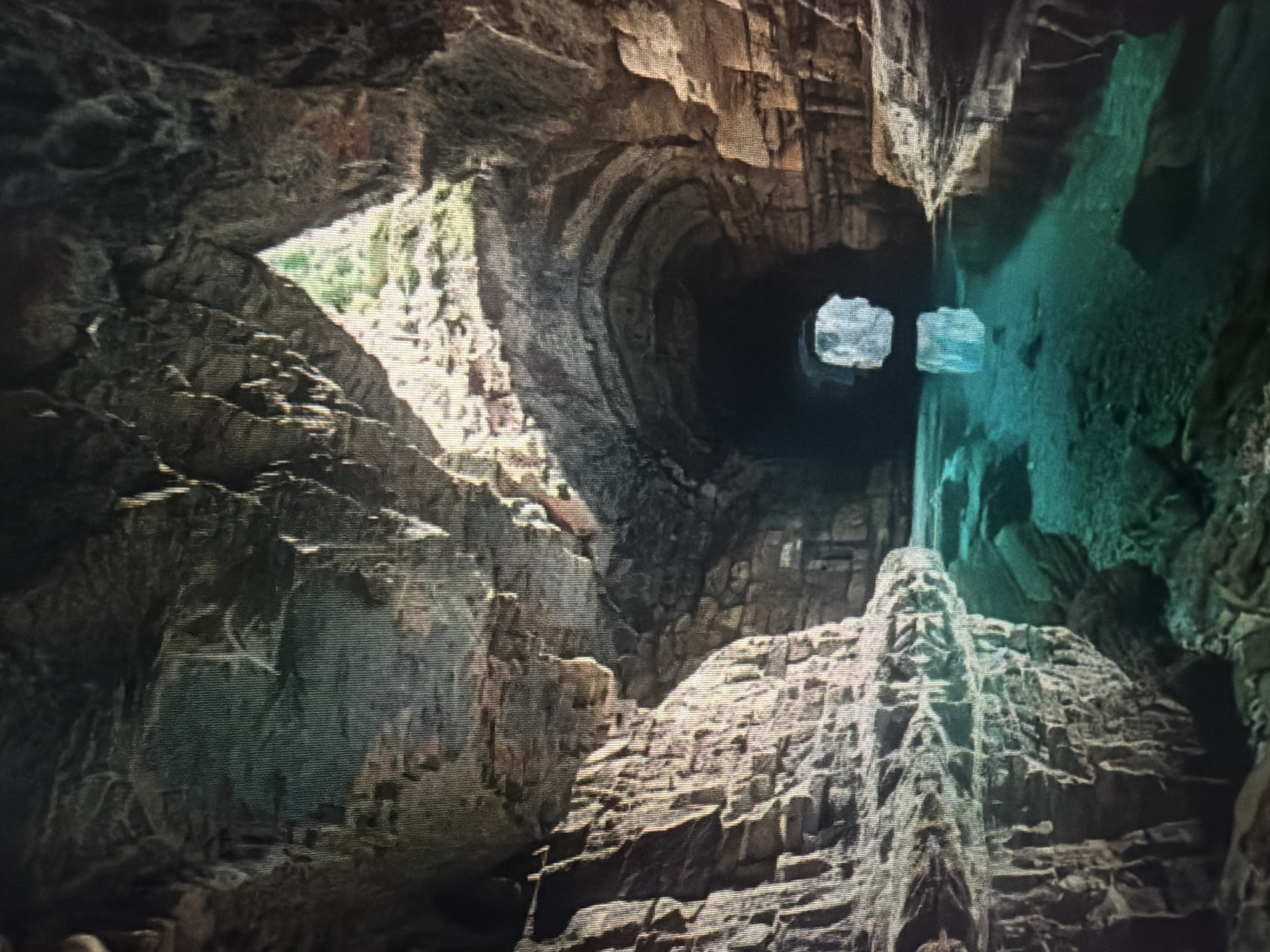 The image shows the interior of a cave with a body of water flowing through it. The cave walls are rough and rocky, with some areas appearing to be made of stacked stones. Light streams in from an opening at the far end of the cave, illuminating the water and the cave's interior. The water is a striking turquoise color and appears to be clear, with the rocky bottom visible in places. The water also reflects the cave's ceiling and walls. The scene suggests a natural geological formation, possibly a sea cave or an underground river. The location in Baan Mae Nam, Thailand, is noted, but the image itself does not contain any specific markers to confirm this location. There are no people or any other discernible subjects or objects in the image besides the natural cave environment and the water.