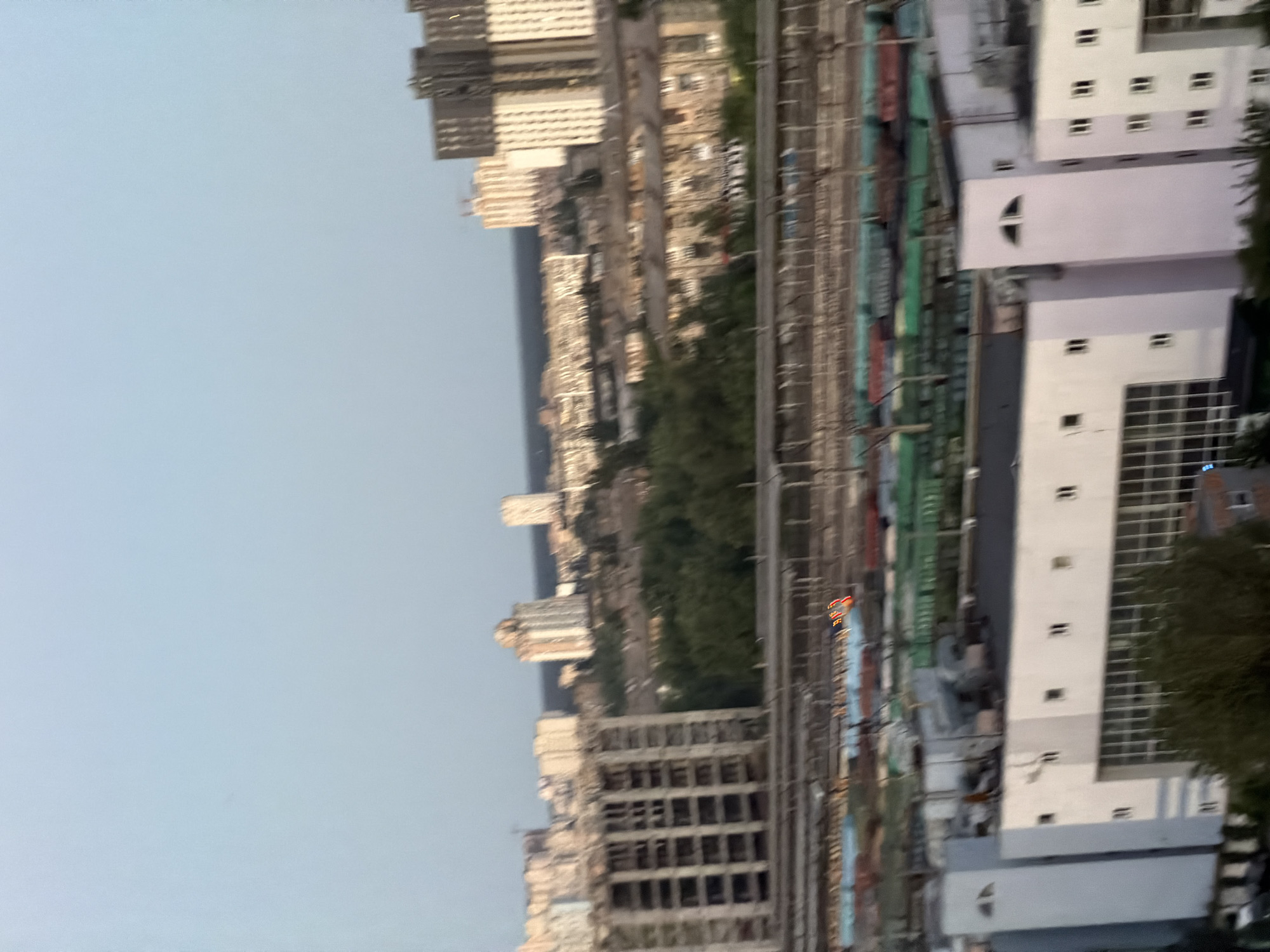 This is an aerial view of an industrial area in Kyiv, Ukraine. In the foreground, there are several low-rise, modern buildings with white facades and large windows. Some of these buildings appear to be industrial or commercial facilities. In the middle ground, there are railway tracks with numerous train cars, some of which are green and red. Beyond the train tracks, there is a dense area of trees, and behind the trees, a skyline of apartment buildings and other structures of varying heights, including some taller, more modern residential towers, are visible. The sky is clear and pale blue, suggesting it is either early morning or late evening. The overall impression is an urban landscape characterized by a mix of industrial infrastructure and residential development.