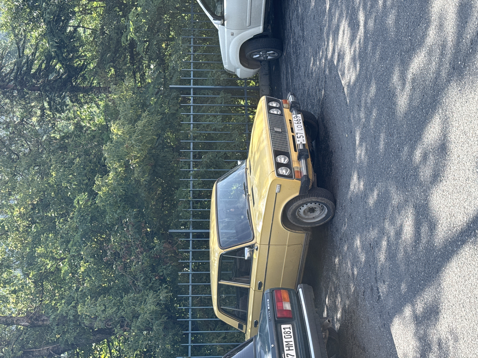 A yellow Lada car and a white SUV are parked on an asphalt surface in front of a metal fence and a dense green tree line. The Lada, a sedan, is positioned to the left of the SUV. The license plate on the Lada reads "57 LO 669 AM". The SUV is a modern crossover model. The scene appears to be outdoors, possibly in a parking lot, with dappled sunlight illuminating the ground. The background consists of lush green foliage.
