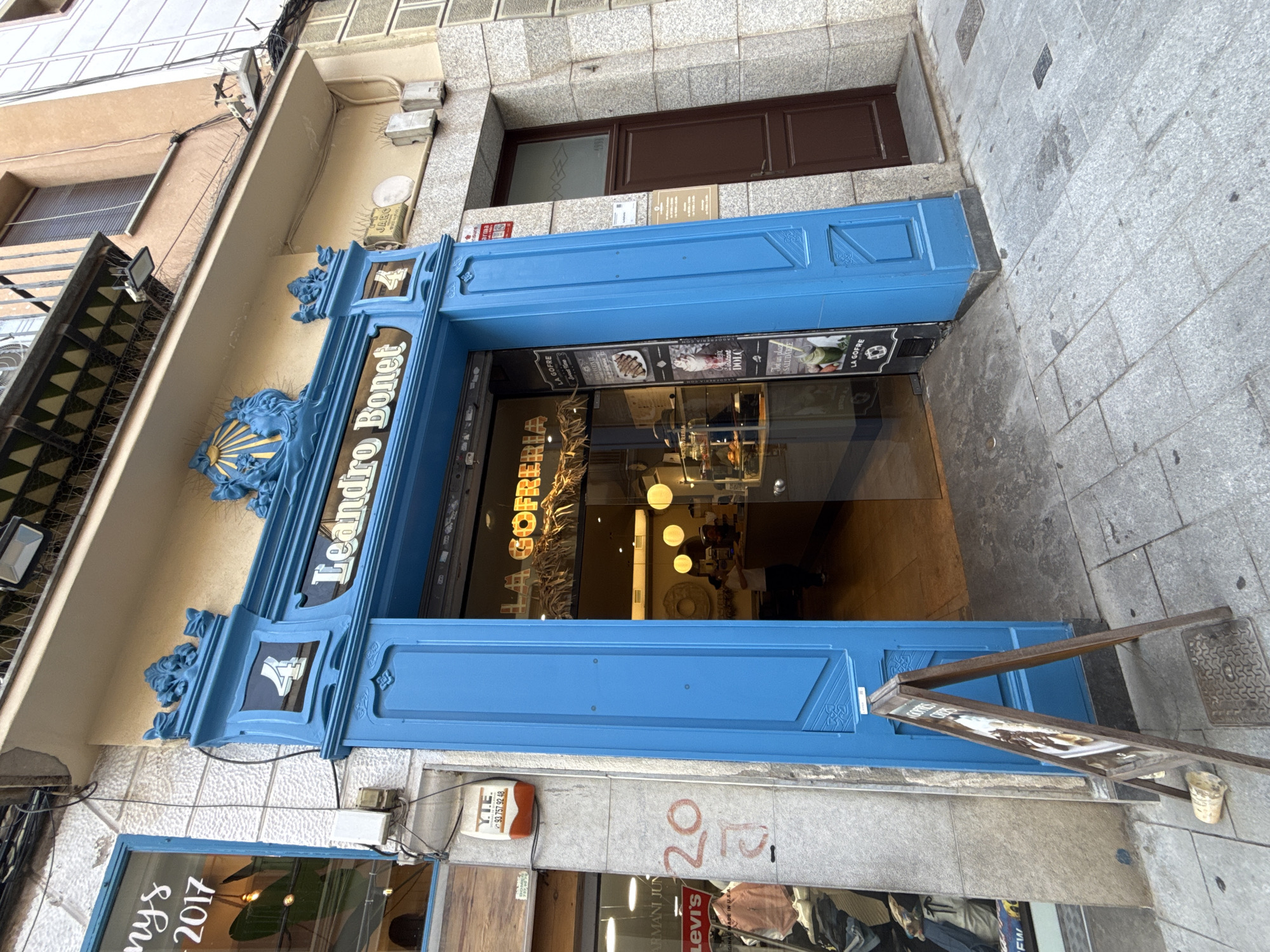 The image shows the exterior of a shop with a bright blue entrance. Above the entrance, the name "Leandro Bonet" is displayed in white lettering. Below this, the words "LA GOFRERIA" are visible in a yellow, stylized font, indicating it is a waffle shop. A sandwich board sign stands on the sidewalk in front of the entrance. To the left of the blue entrance, a store window displays clothing and the year "2017". To the right of the blue entrance is a brown door. The shop is located on a paved street. A person is visible inside the shop, standing behind a counter. The building facade is painted a light yellow and textured grey.