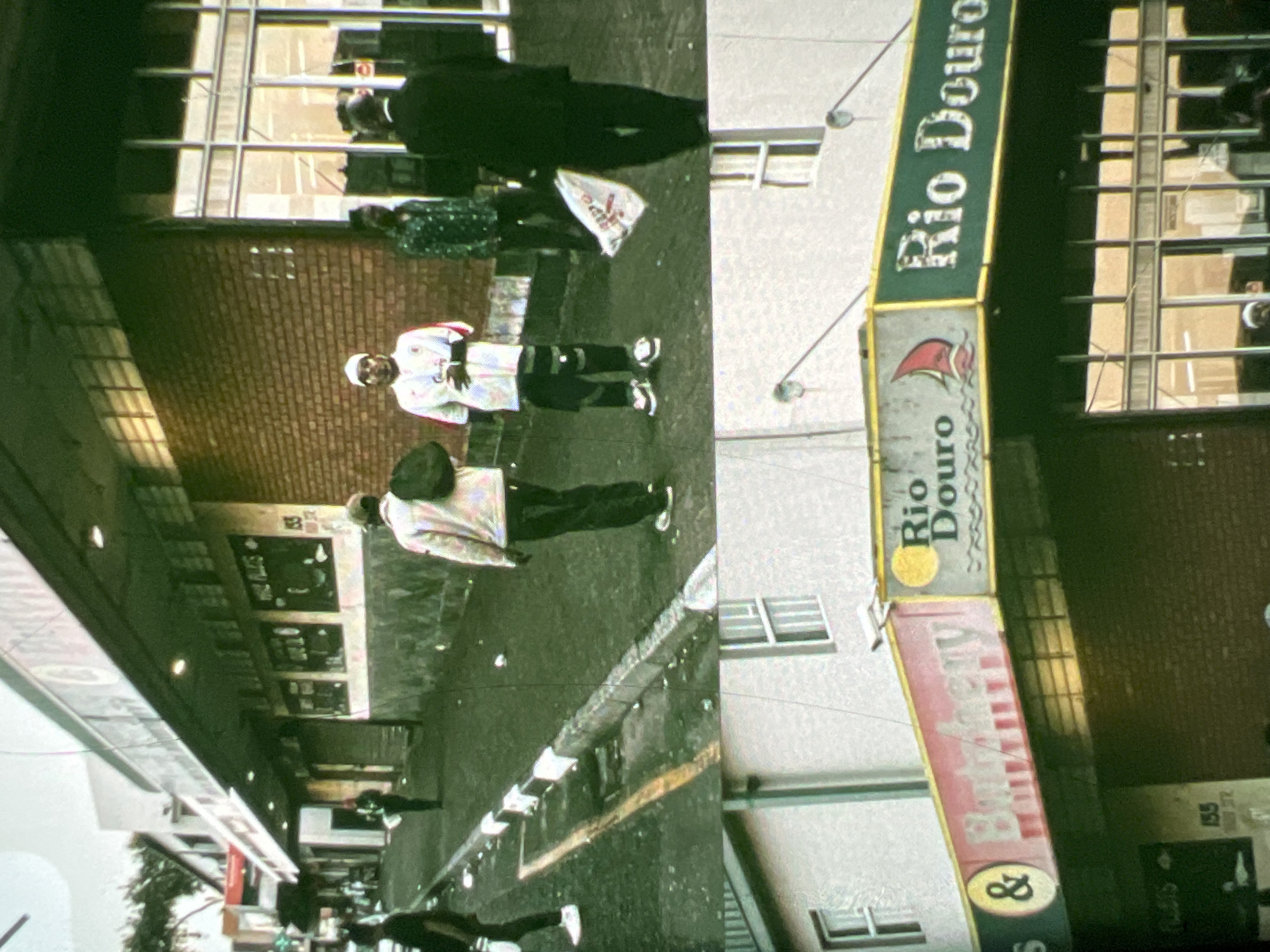 The image shows a street scene in Lagos, Nigeria, featuring a row of shops. In the upper section, a group of people are visible walking on a street lined with buildings and storefronts. One person is wearing a white and red jersey, another a patterned green shirt, and a third a dark suit. A white flag with red lettering is on the ground. The lower section of the image focuses on a shop with signs that read "Rio Douro" and "& Butchery". The shop has a green awning and large glass windows. The scene appears to be a commercial area with pedestrian traffic.