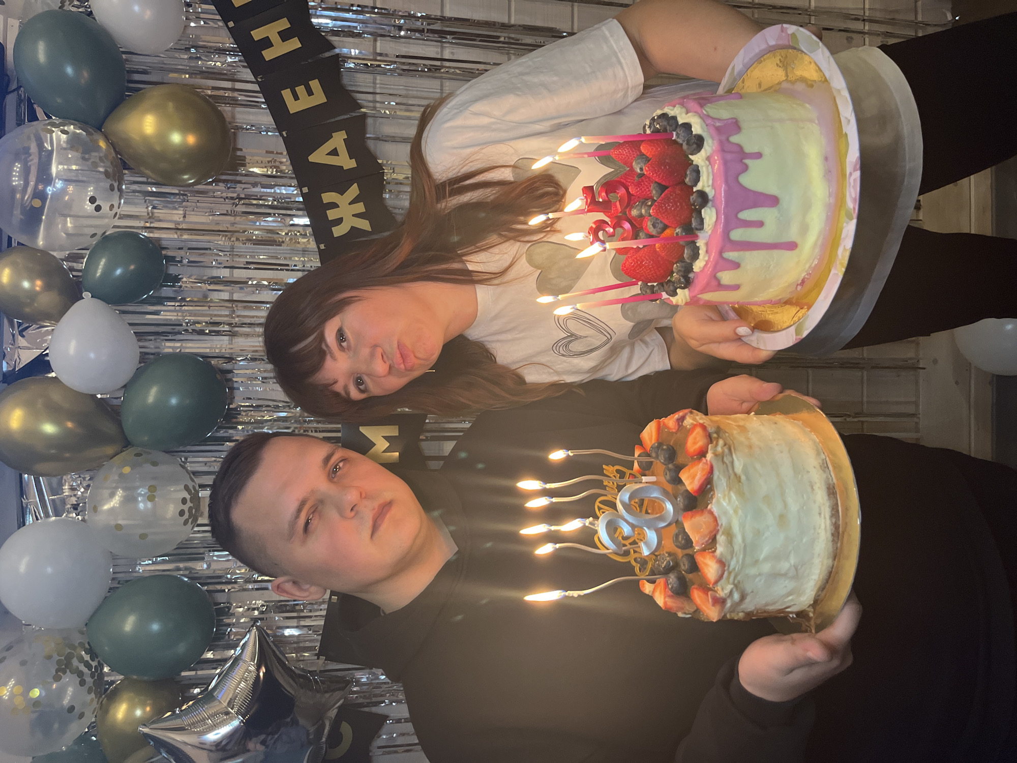 The image shows a man and a woman holding birthday cakes in an indoor setting. The man, on the left, is wearing a black hoodie and holds a cake with "30" candles and strawberries and blueberries. The woman, on the right, is wearing a white t-shirt with heart designs and holds a cake with "35" candles and strawberries and blueberries, decorated with pink icing. The background features silver streamers and a balloon display, including white, gold, silver, and green balloons. A black banner with gold lettering is also visible. The location is undetermined.