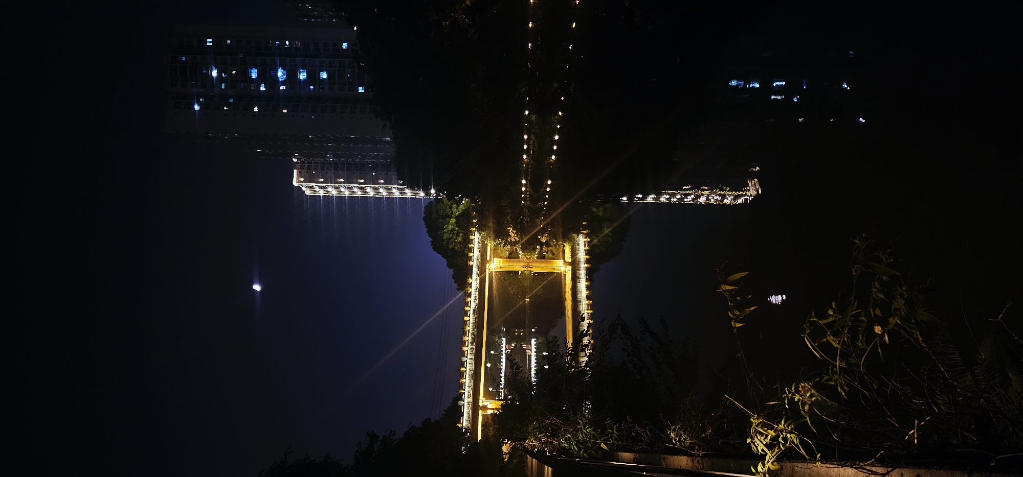 The image depicts a nighttime scene in Bishan, China. A body of water, likely a river or canal, occupies the lower half of the frame, reflecting the lights from the surrounding environment. A bridge, illuminated with warm yellow lights, spans across the water in the middle ground. On the right side of the image, tall, modern buildings are visible, their windows aglow with blue and white light. The sky above is dark, with a crescent moon visible towards the upper left quadrant. Trees and vegetation line the banks of the water and partially obscure the view of the buildings. The overall impression is of an urban landscape at night, with artificial lighting creating a vibrant contrast against the dark sky and water.