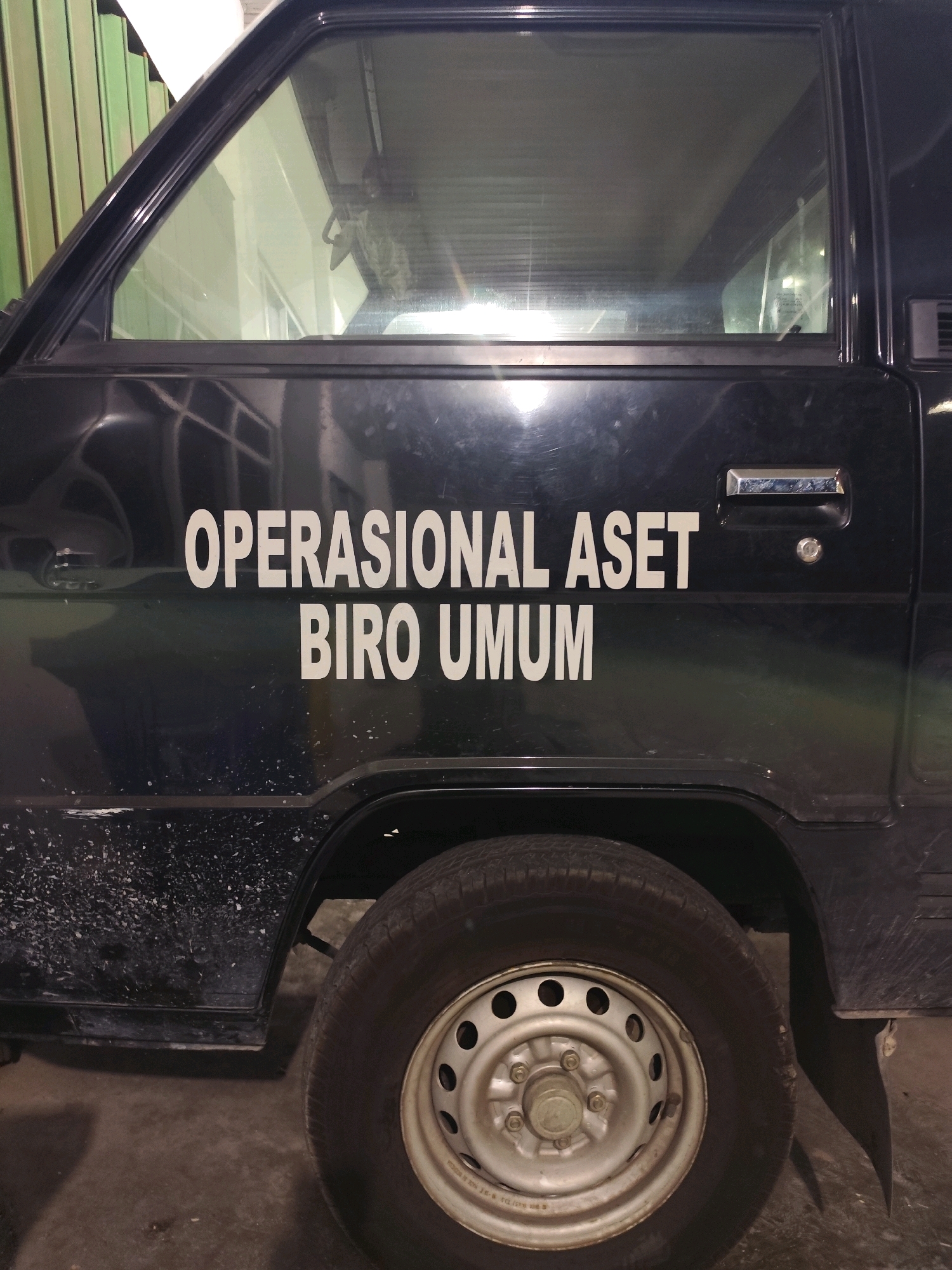 A black van is parked indoors, possibly in a garage. White text on the side of the van reads "OPERASIONAL ASET BIRO UMUM". The van is dirty, with visible water spots and dust on its exterior. A silver wheel with lug nuts is visible on the right side of the van. The background shows a metallic green structure and a light-colored wall. The scene is not indicative of any specific location within the city of Palangka Raya, Indonesia.